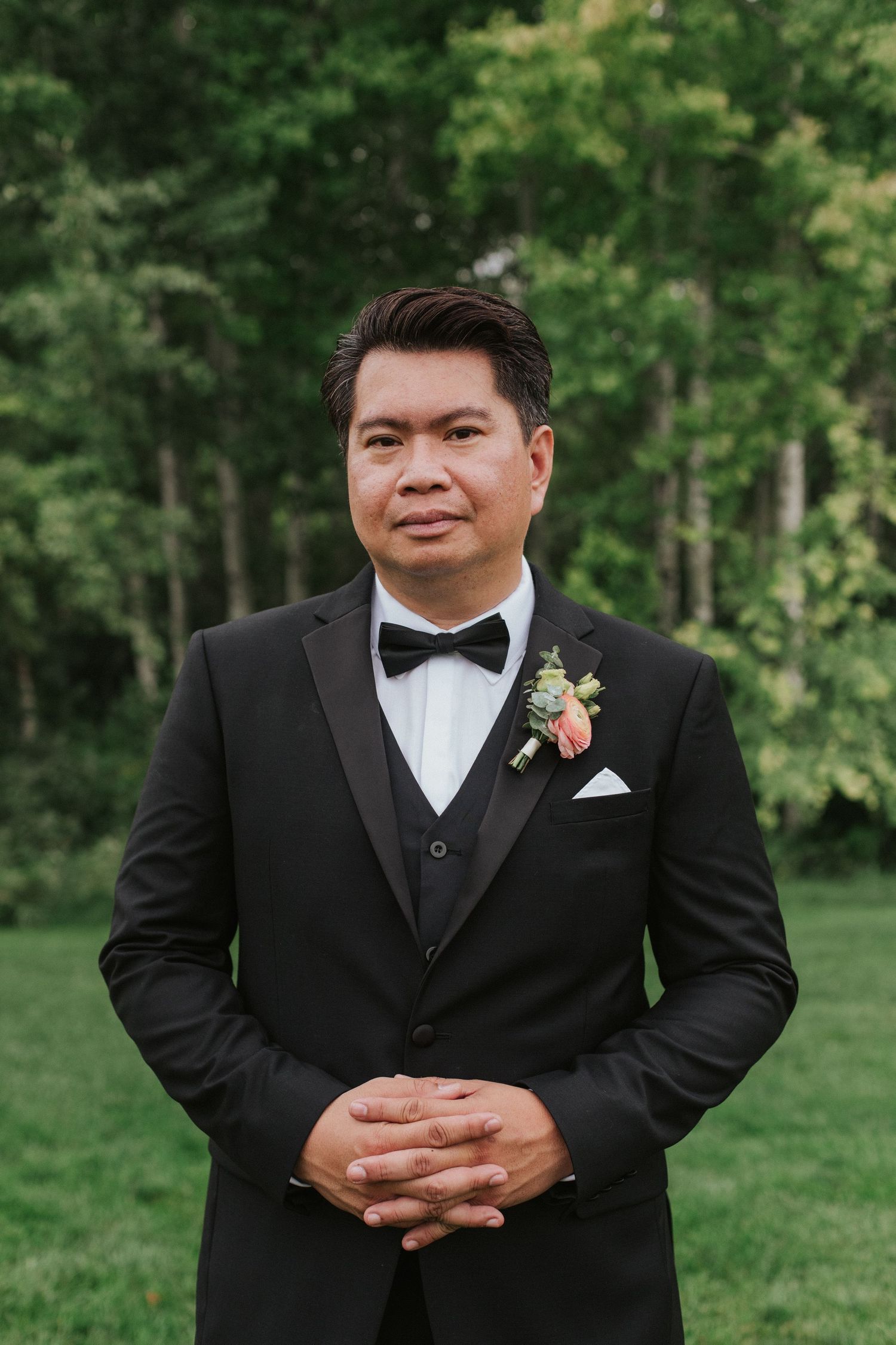 A groom in a classic black tuxedo and bowtie poses for formal portraits in a lush outdoor garden setting.