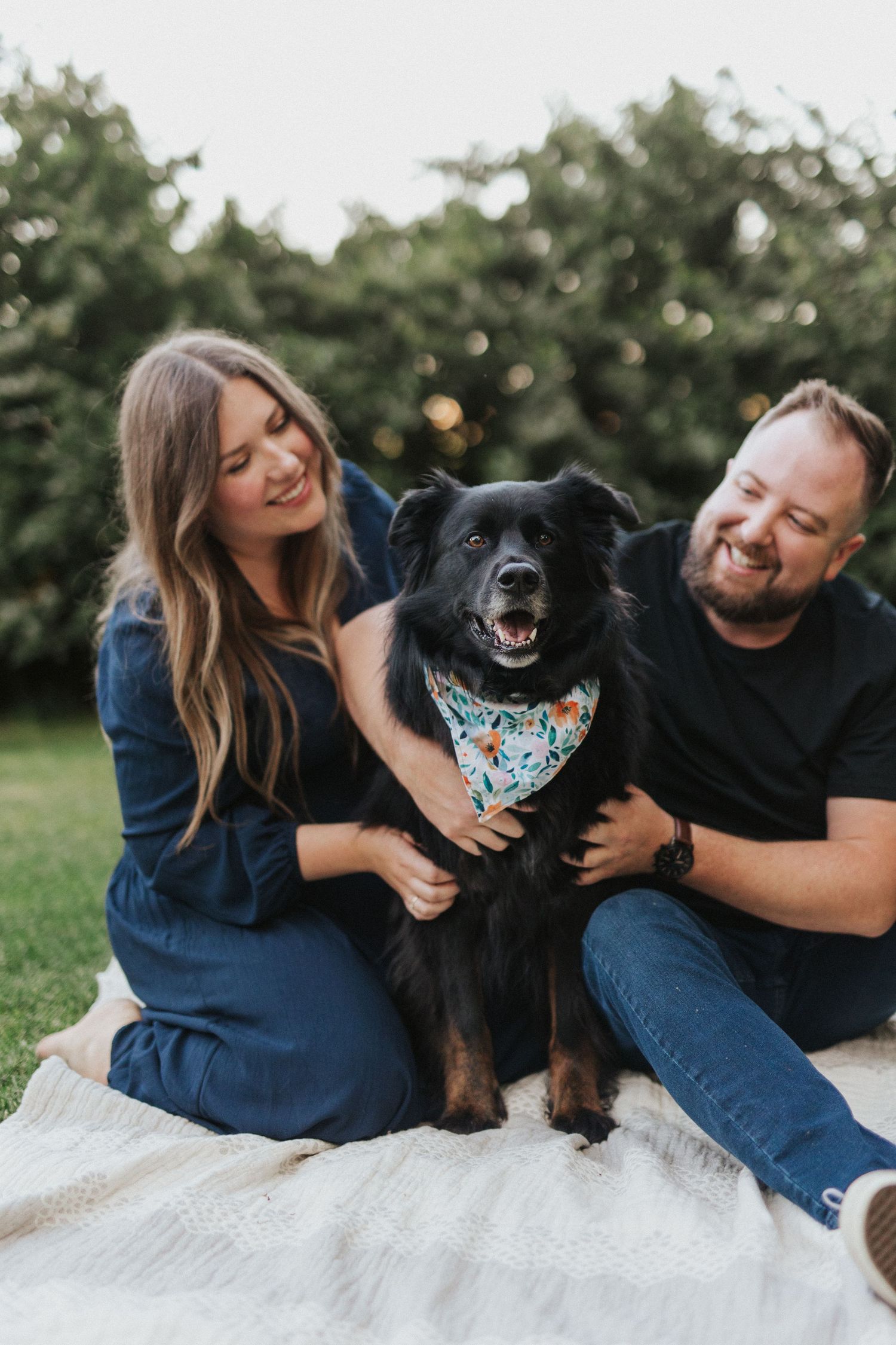 Happy couple lounges on blanket with their black dog wearing a floral bandana.