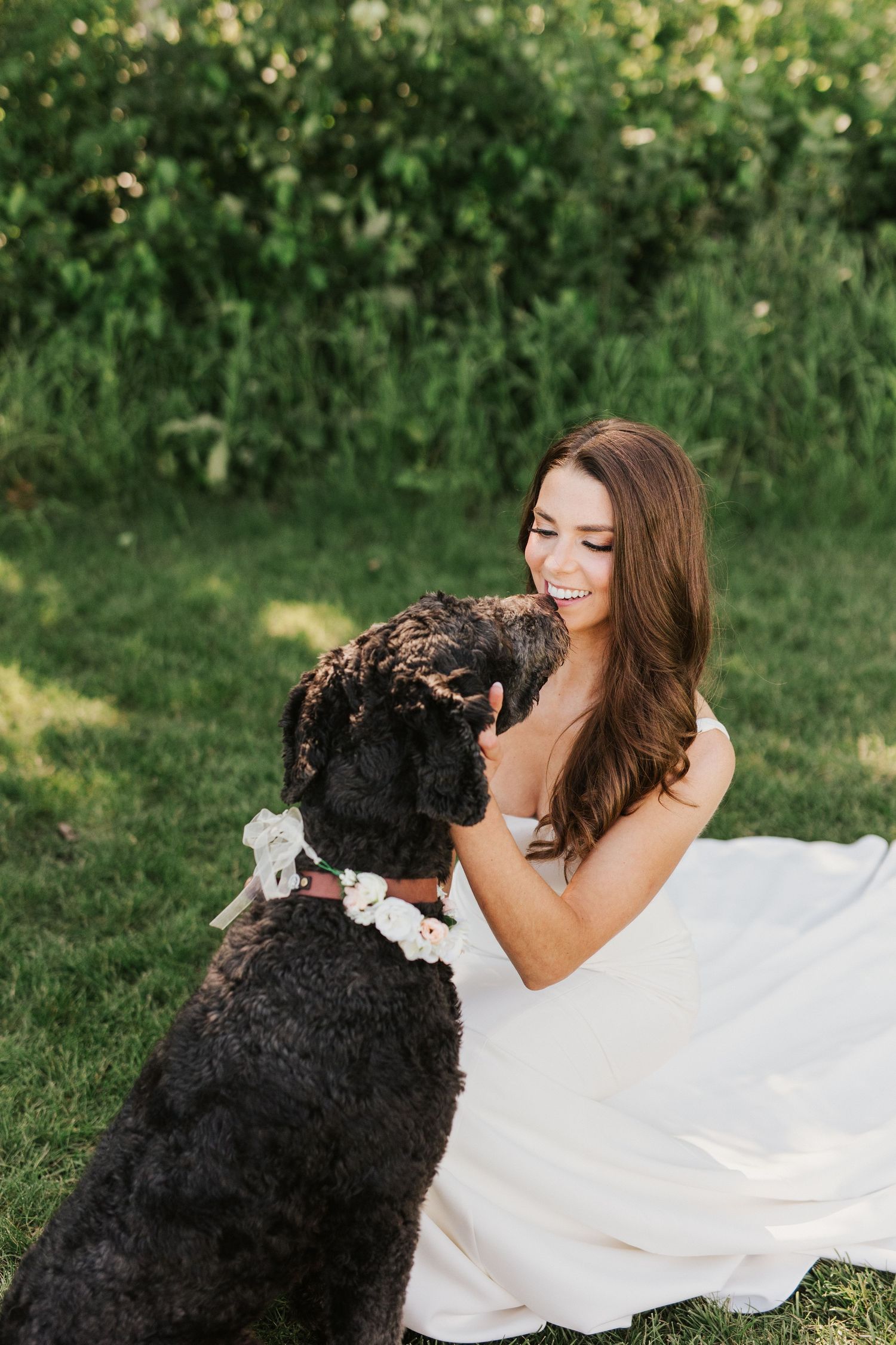 Bride in white dress sharing a tender moment with black dog on grassy lawn.