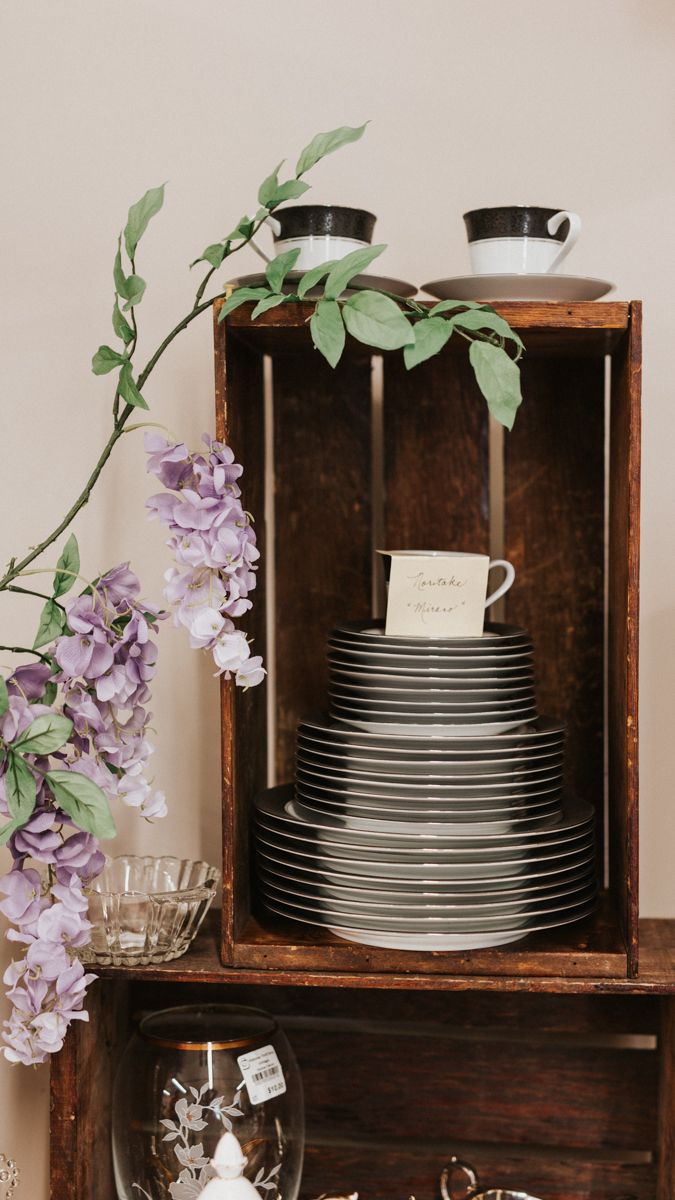 Rustic wooden shelving unit displaying stacked white plates and purple flowers.
