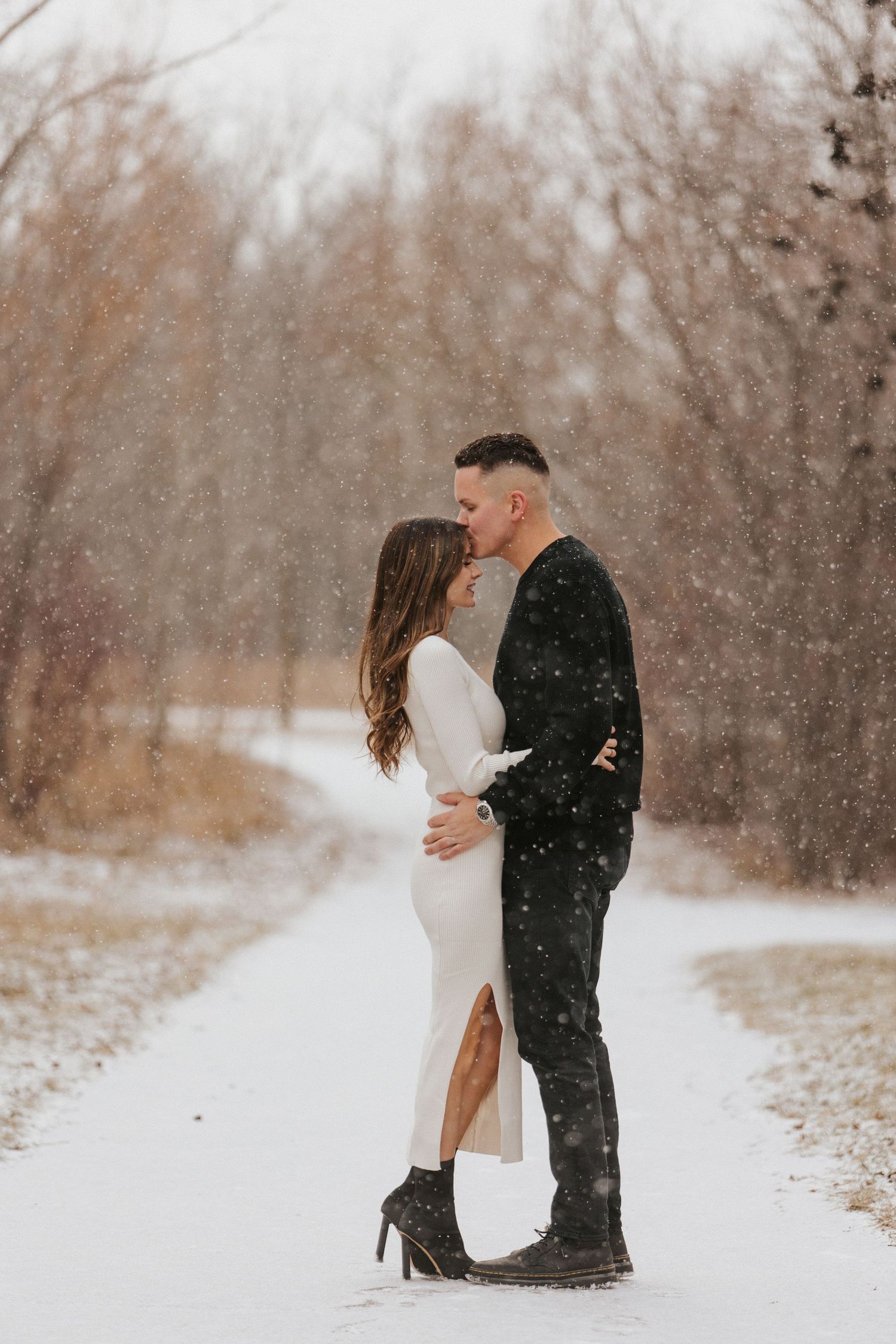 A romantic winter scene with two people embracing in falling snow surrounded by bare trees.