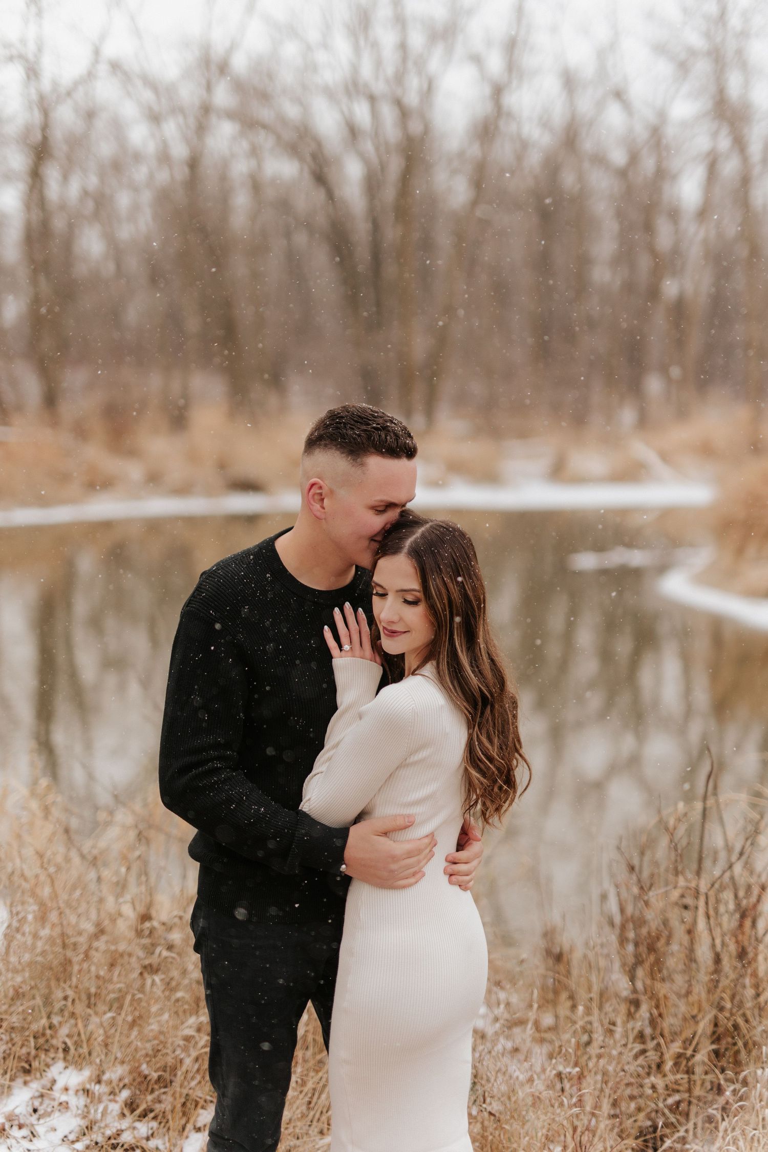 A couple shares an intimate moment by a snow-covered lake with bare winter trees in the background.