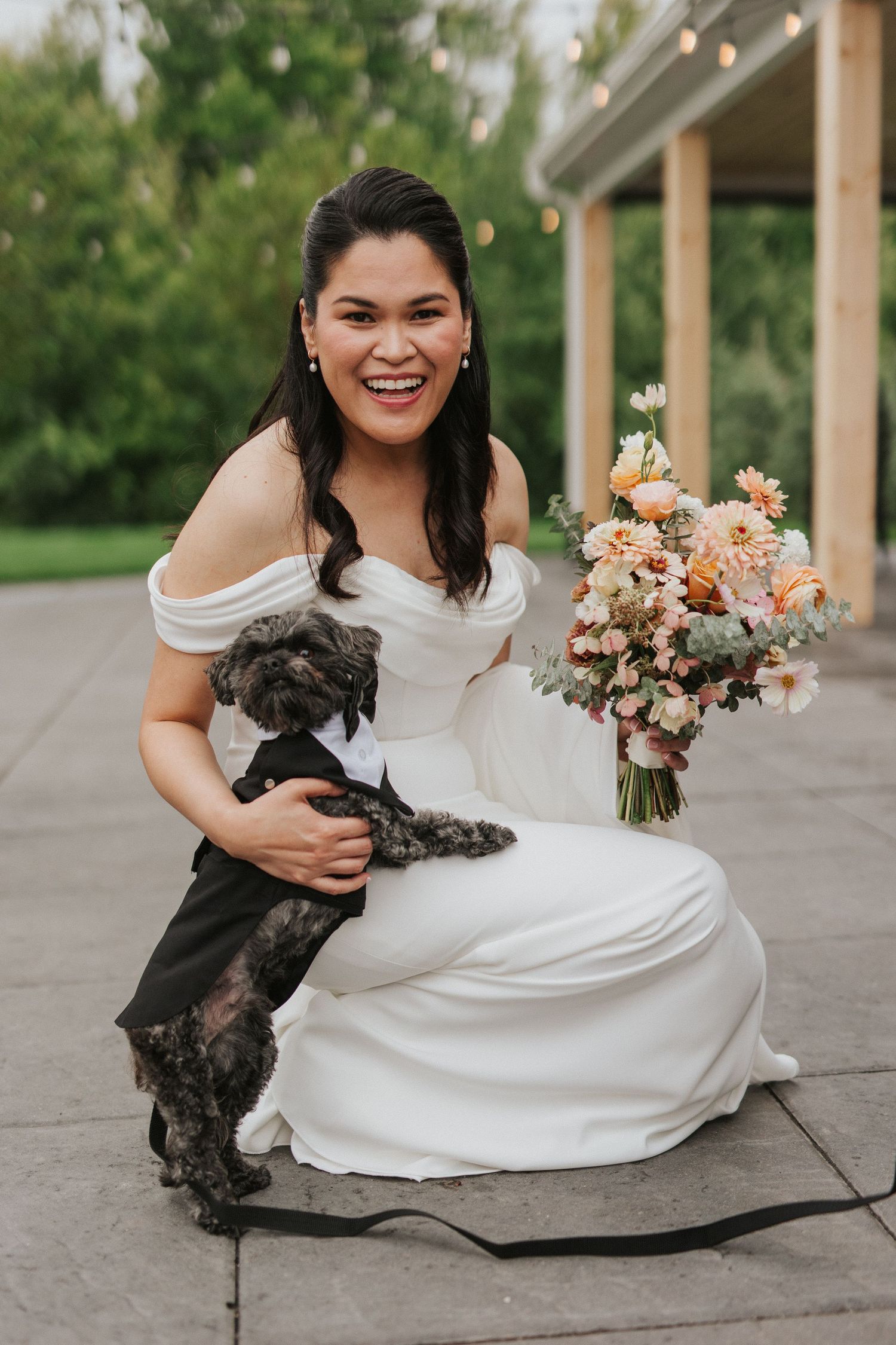 A bride in an off-shoulder white dress kneels while holding a small dark dog and peach-colored floral bouquet.