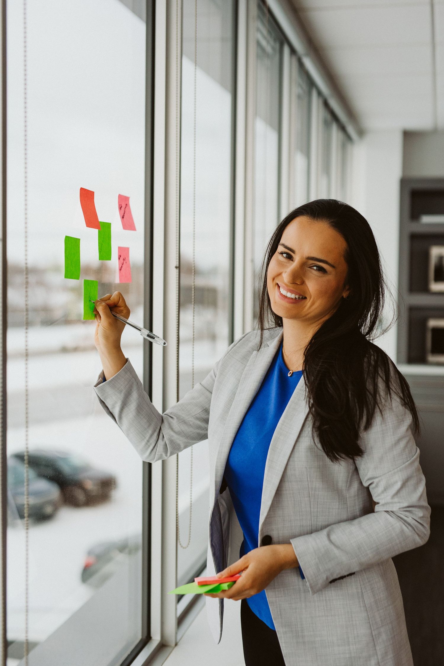 Professional dressed in a gray blazer and blue top holds colorful sticky notes by office windows.