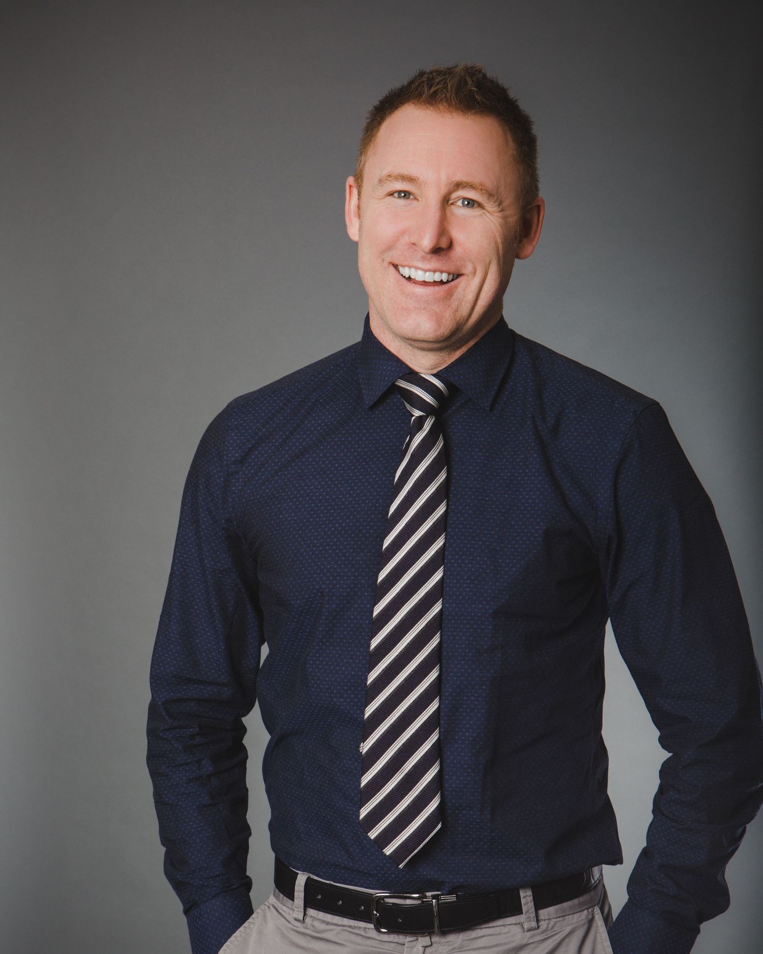 Professional corporate portrait in navy suit and striped tie against grey background.