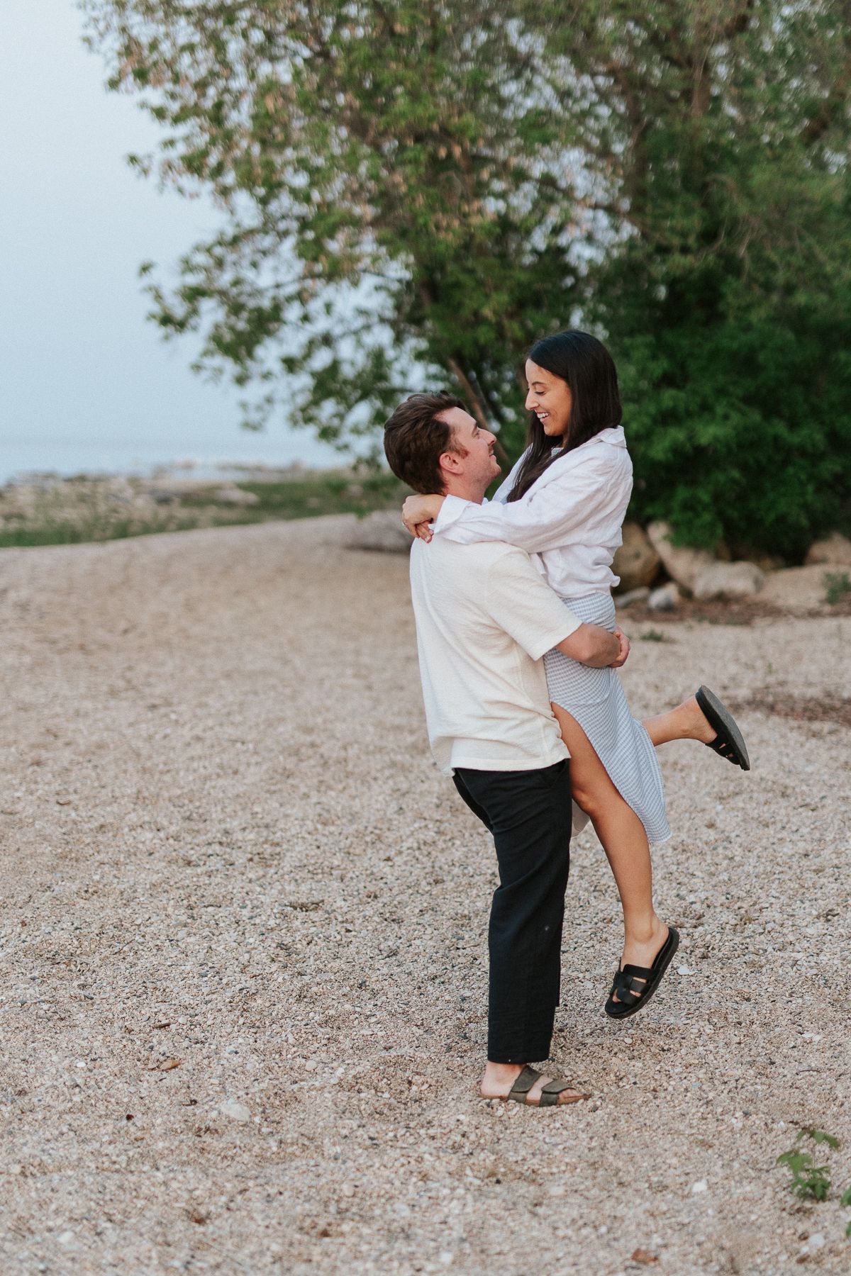 Gimli Engagement photography. Playful couple dancing and twirling on a sandy beach near trees.