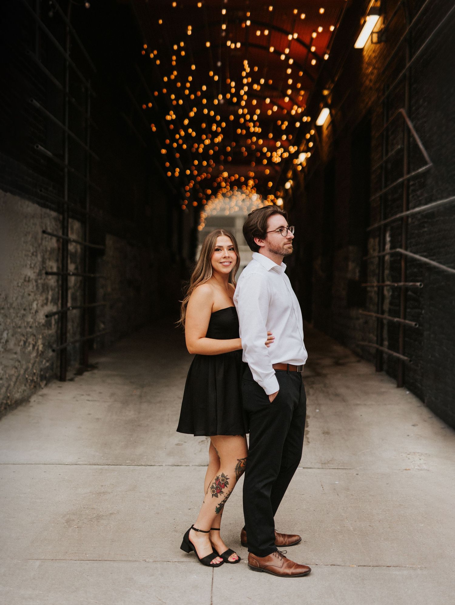 Romantic couple pose beneath twinkling string lights in an urban alleyway.