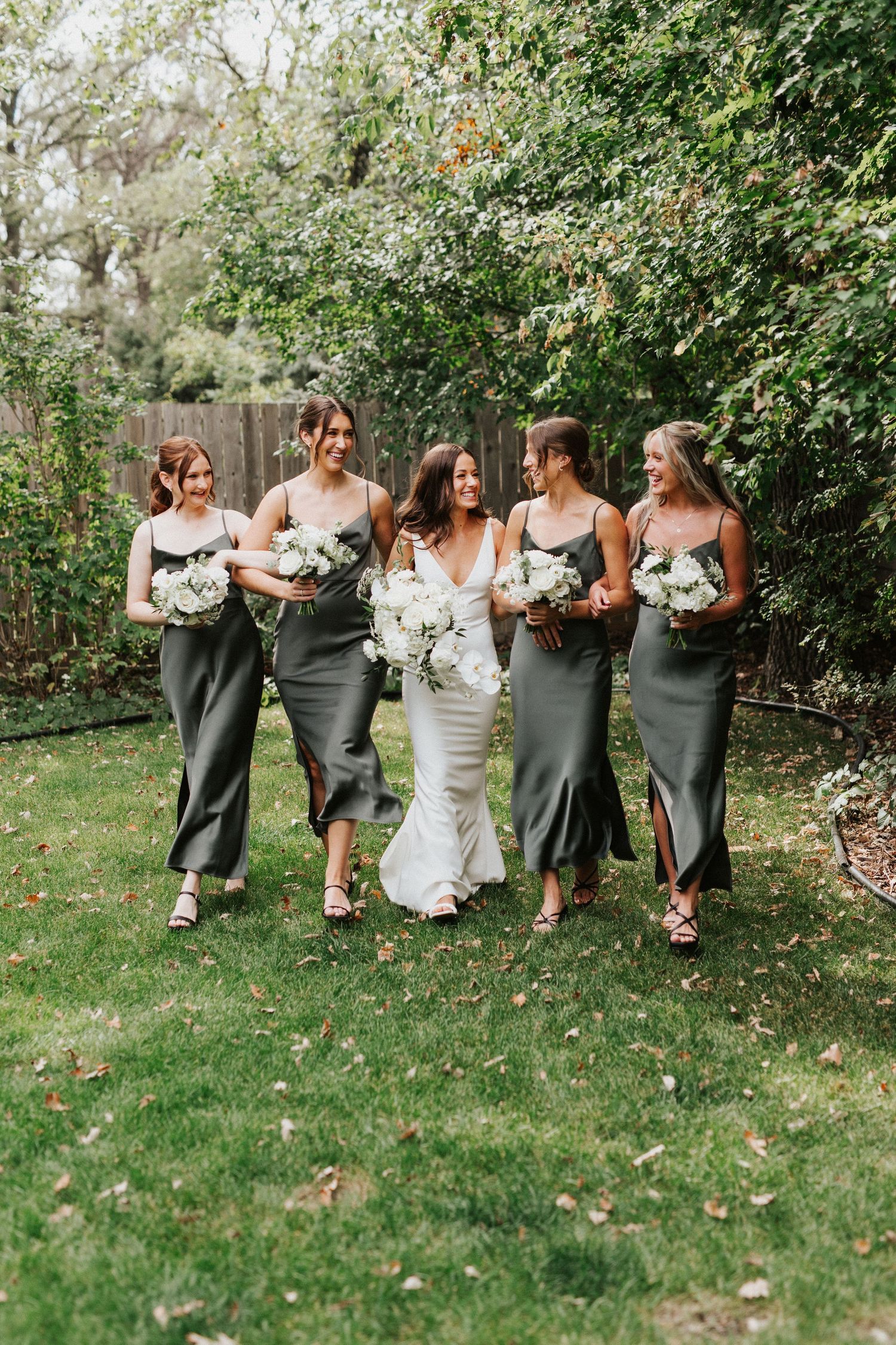 Bridesmaids in sage green dresses walk together through garden setting.