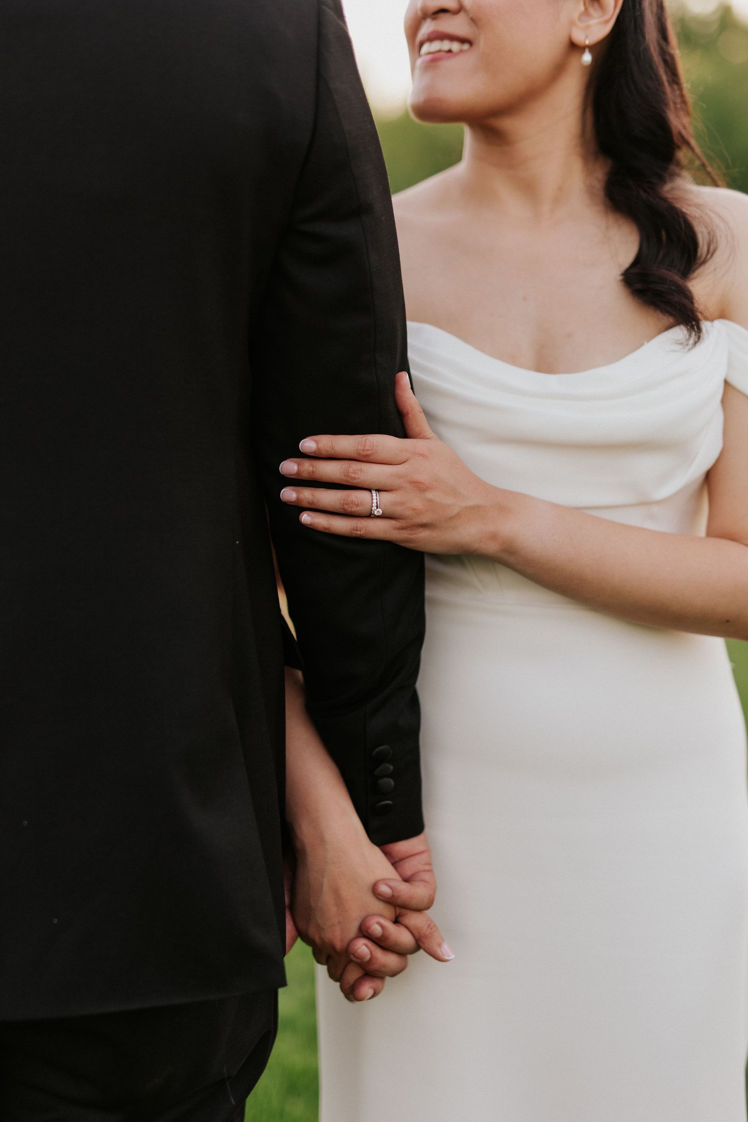 Close-up detail of bride in white dress holding groom's hand and showing wedding ring.