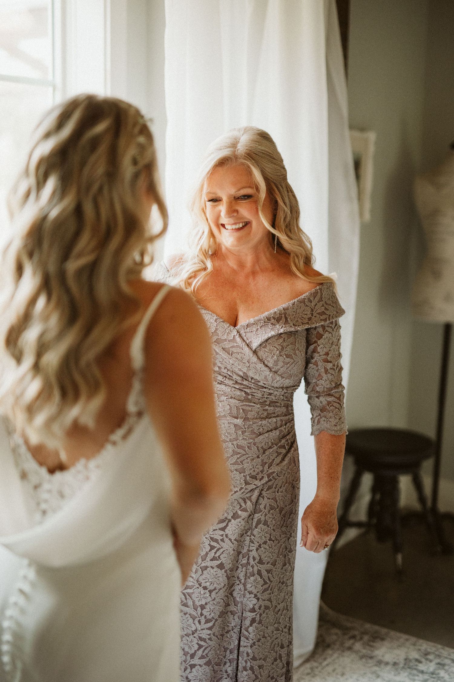 Two women in elegant dresses embrace during wedding preparations.