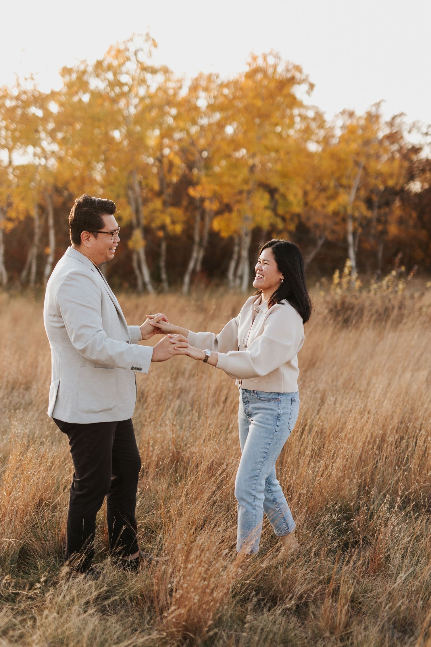 Couple sharing joyful moment in tall autumn grass with golden trees in background.