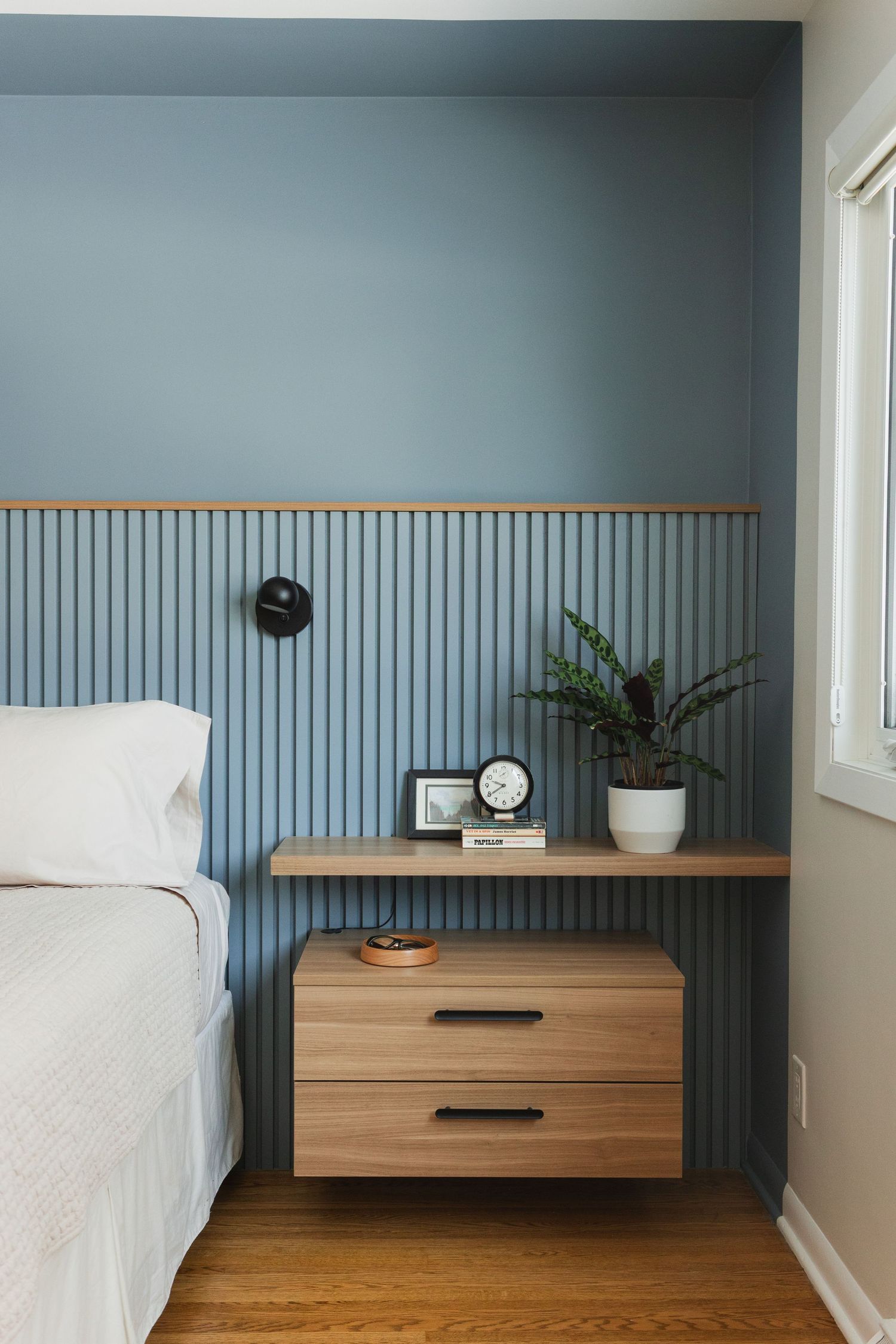 Modern bedroom features a sleek wooden floating nightstand against a blue-gray wall with vertical wainscoting detail.