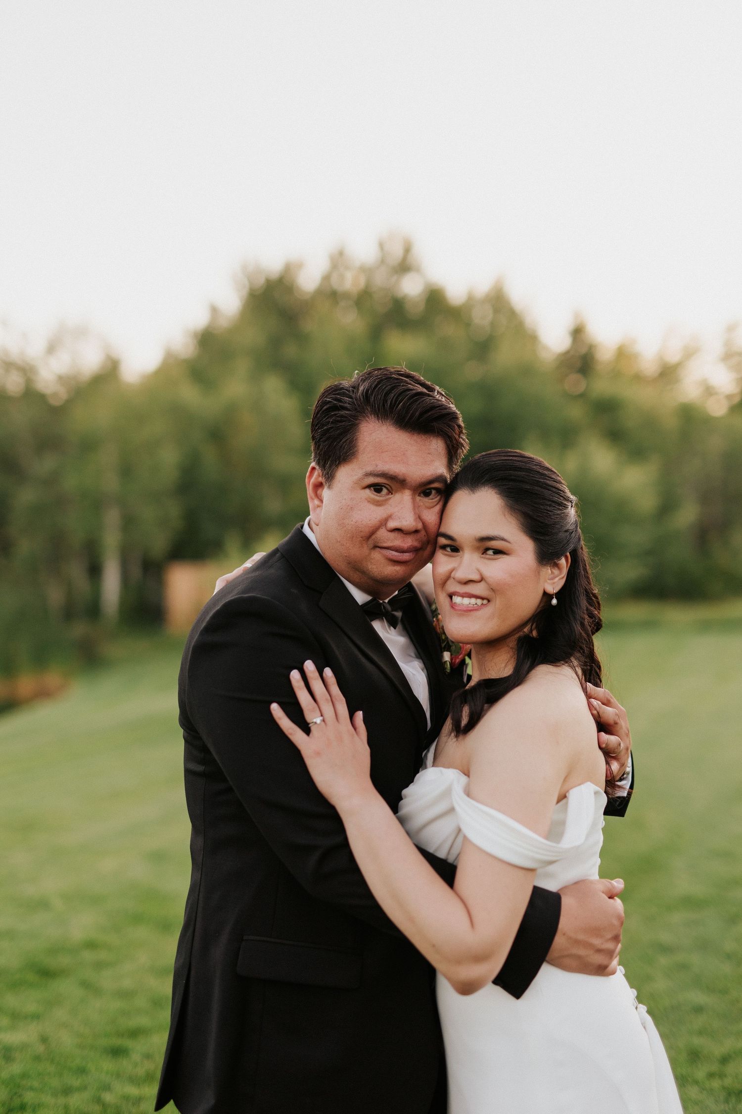 A romantic wedding portrait shows a couple embracing in a sunlit field with lush greenery behind them.