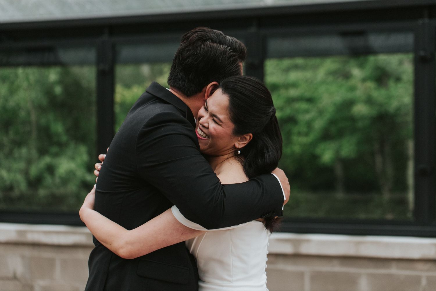 A romantic couple in black and white formal attire embrace during what appears to be a wedding celebration outdoors.