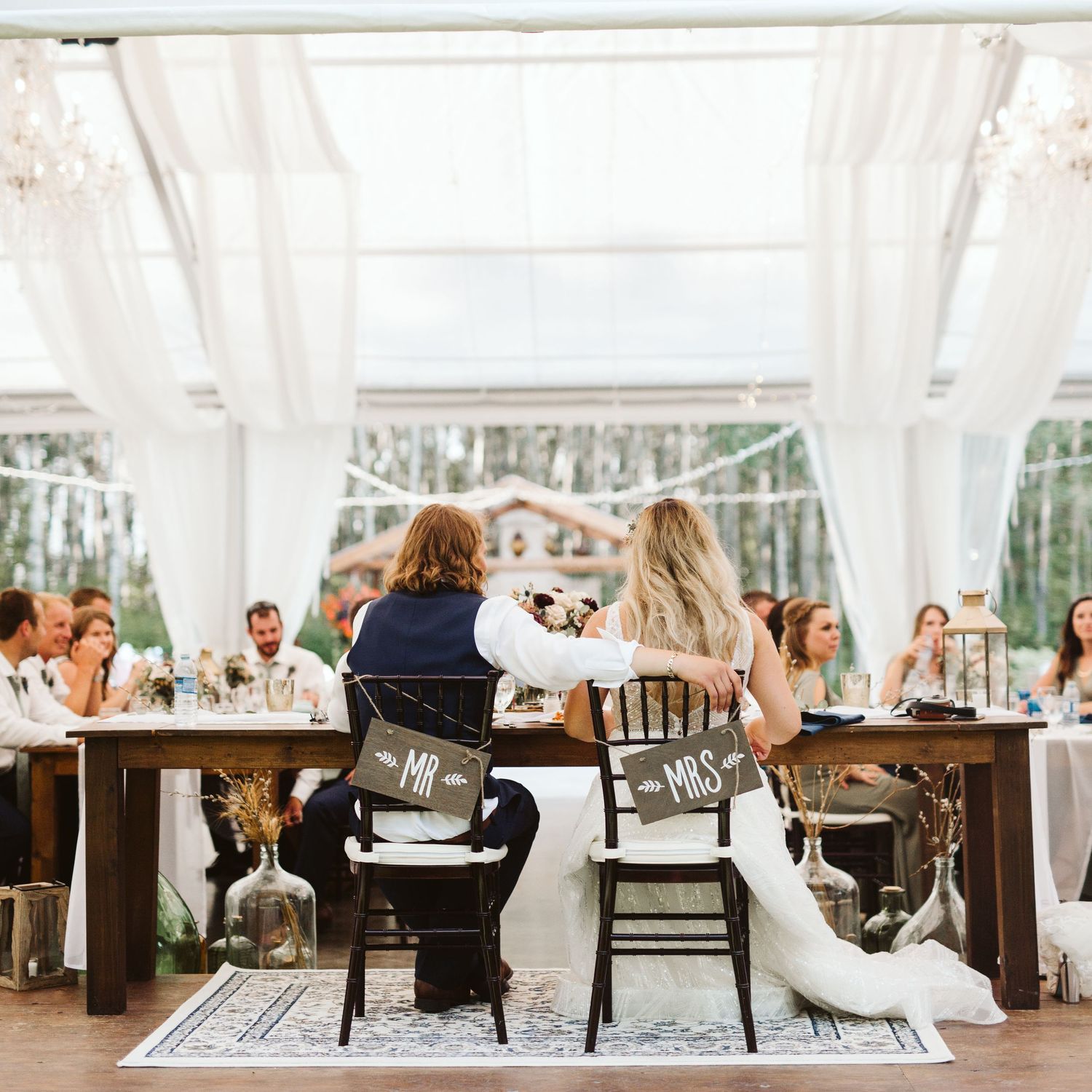 Wedding guests enjoy an elegant outdoor reception with white drapes and rustic wooden furniture.