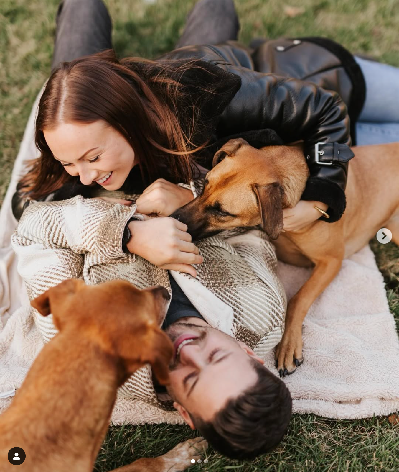Couple lying on blanket getting kisses from their brown dog in the grass.