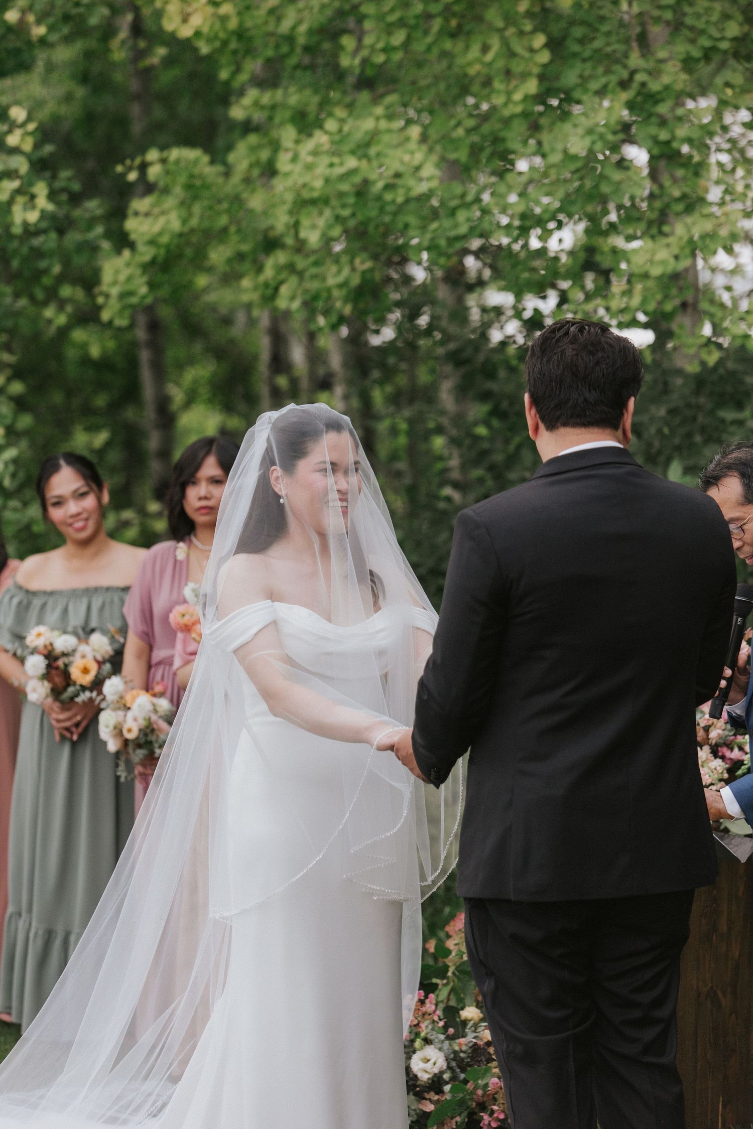 An outdoor wedding ceremony with guests seated in chairs among green trees and natural scenery.