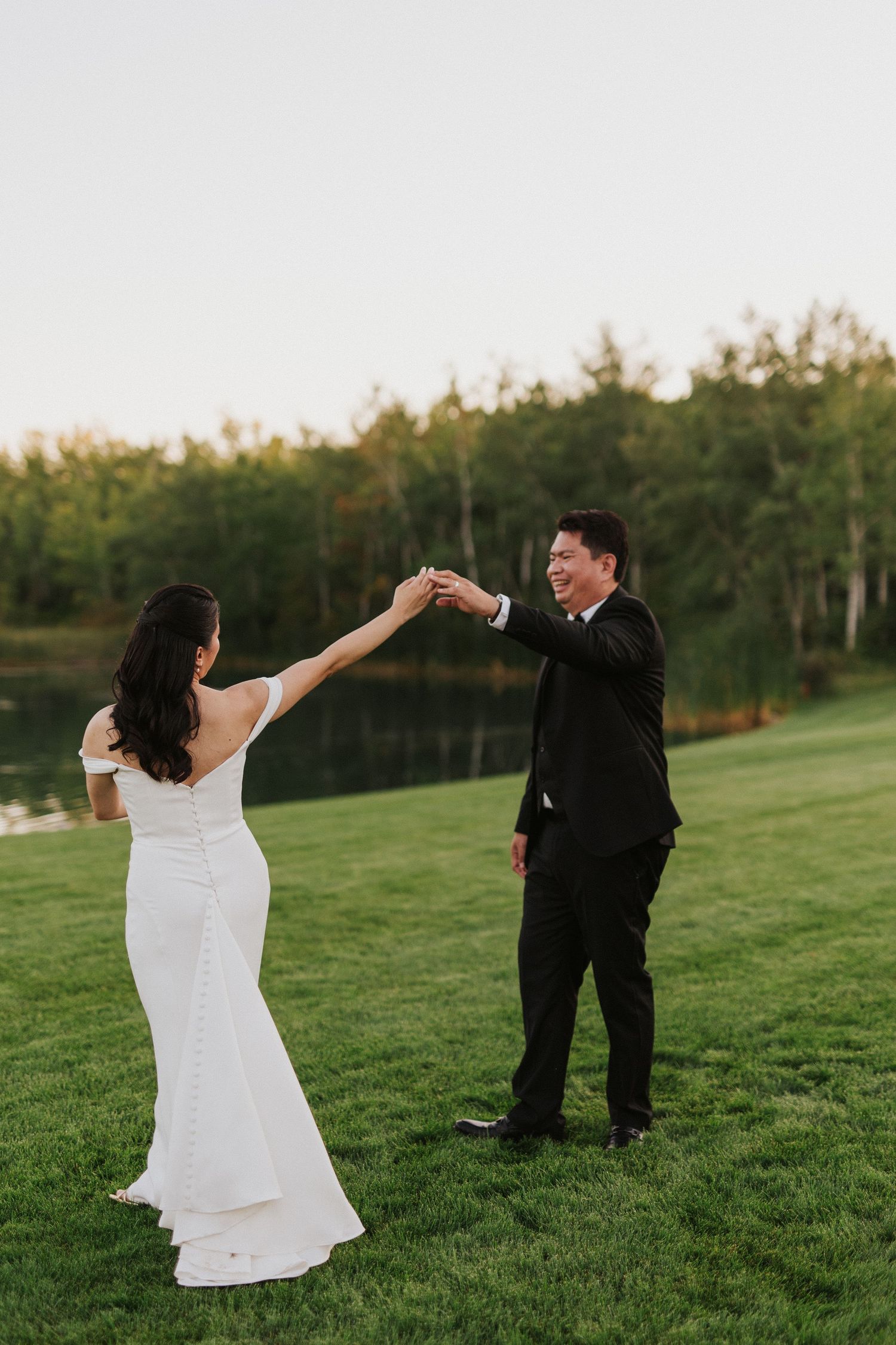 A couple twirling and dancing playfully on a grassy hill with forest backdrop.