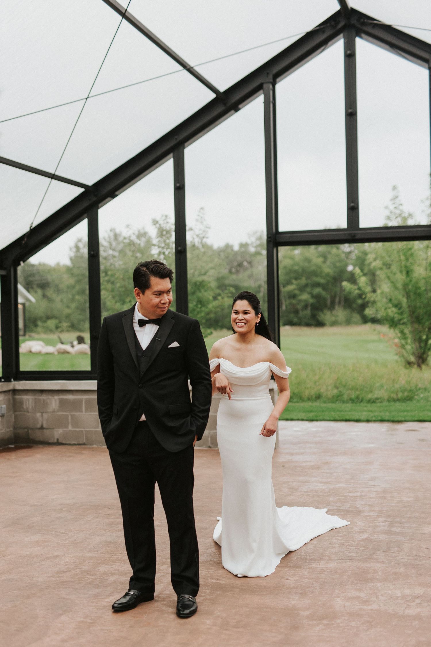 A couple in wedding attire shares a playful moment in a greenhouse venue with glass walls and brick details.