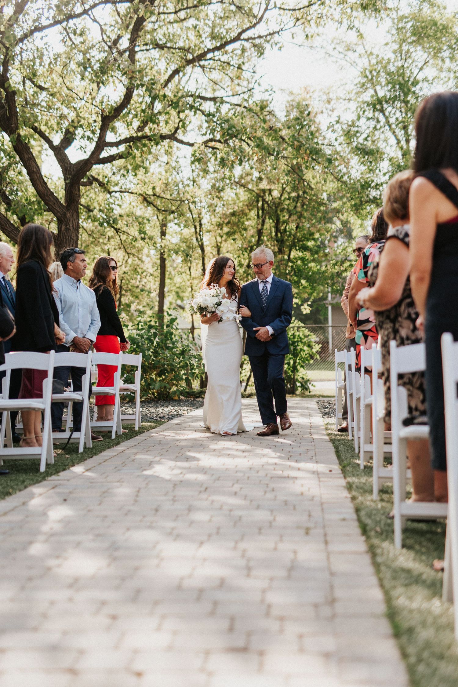 Tree-lined wedding ceremony aisle with white chairs and guests in attendance.