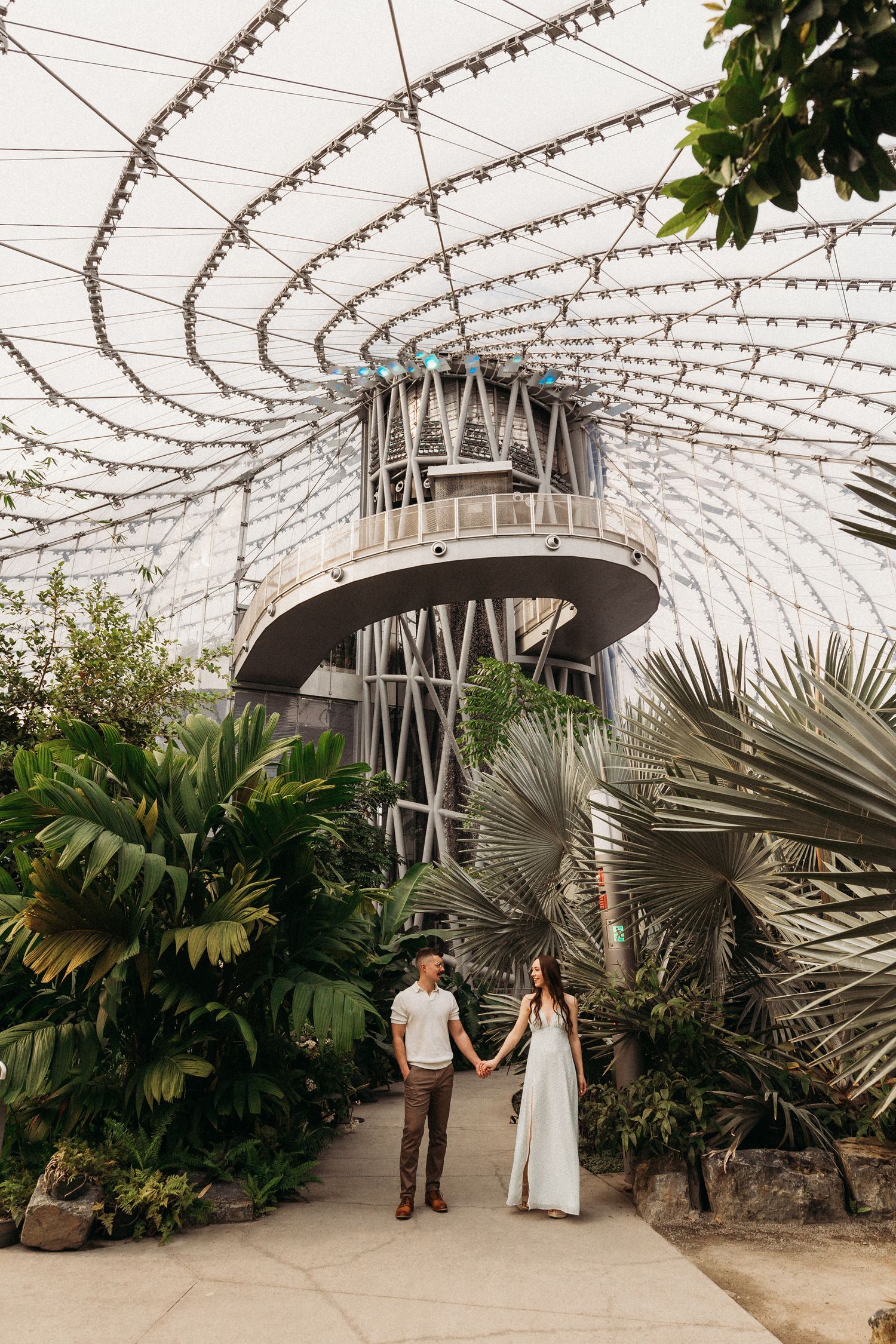 Couple walks hand in hand through a large greenhouse filled with tropical plants and string lights.