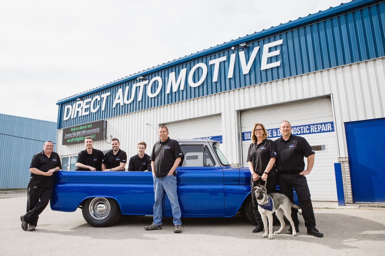 Panoramic view of blue automotive repair shop exterior with vehicles and staff.