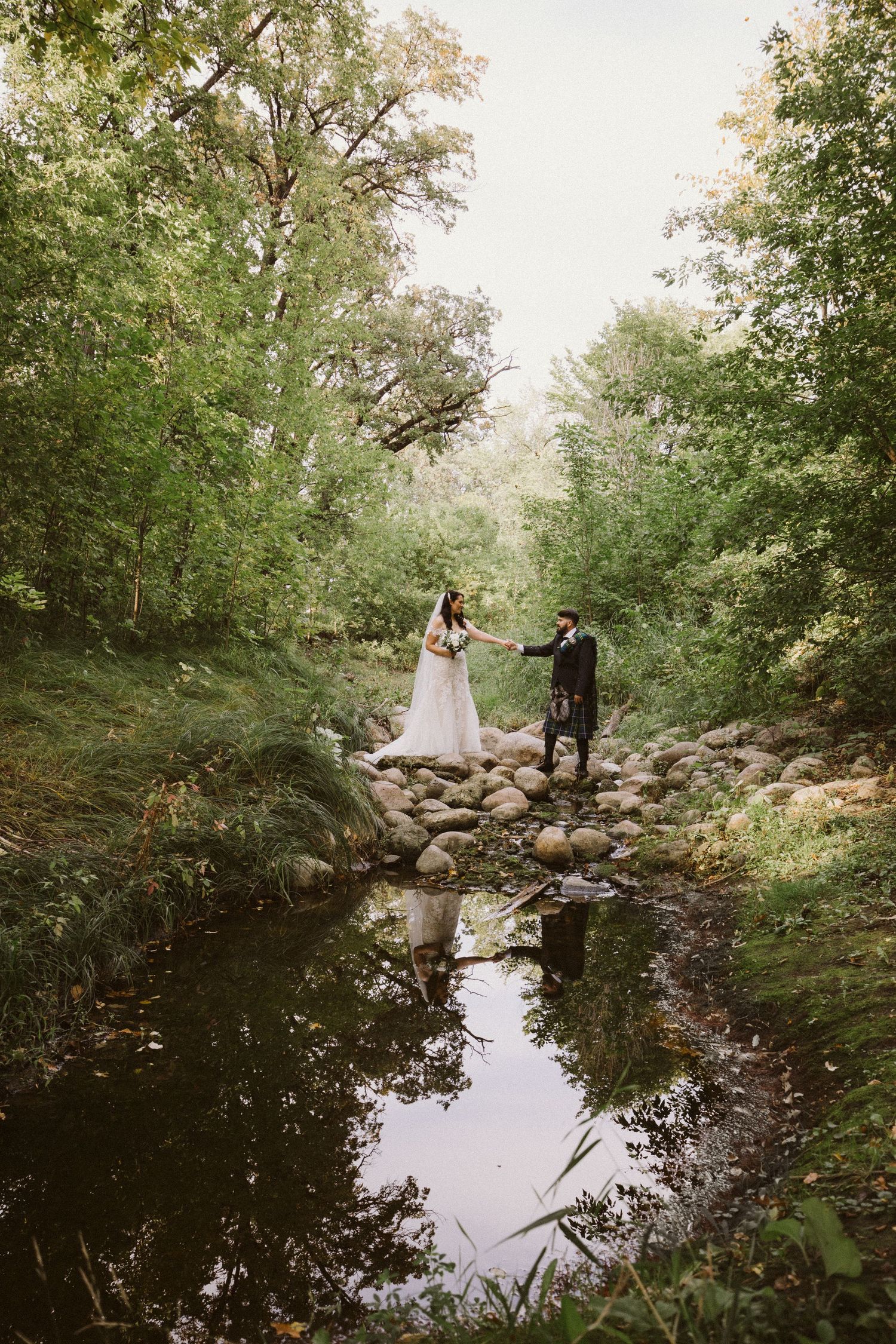 Wedding couple walking near tranquil pond surrounded by lush greenery.