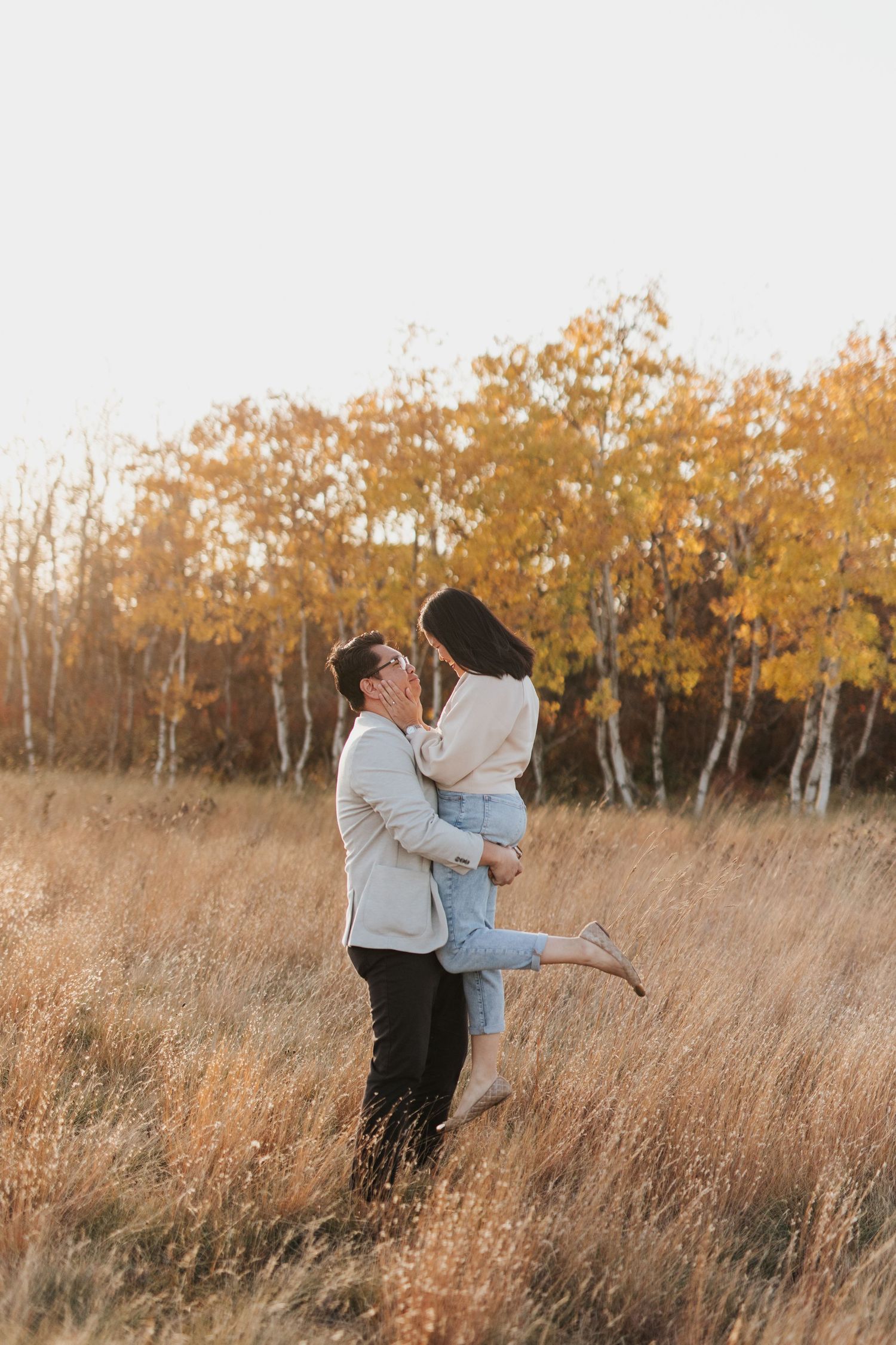 Couple embraces in a golden autumn field with birch trees in the background.