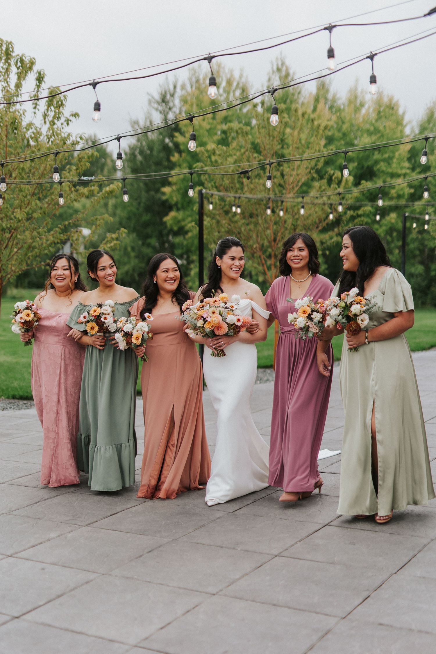 Wedding party in coordinated dresses in shades of pink, sage, and cream hold floral bouquets under string lights outdoors.