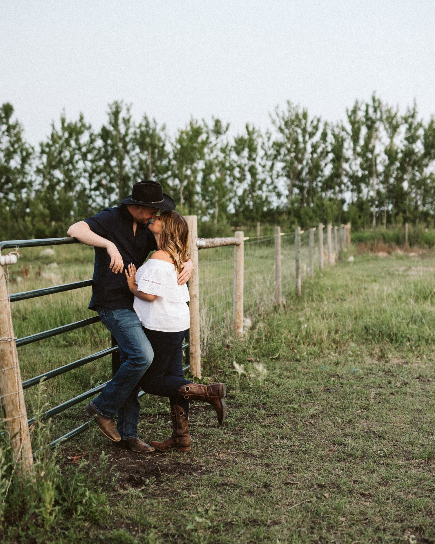 A romantic couple embraces on a grassy hillside with wooden fence posts at sunset.