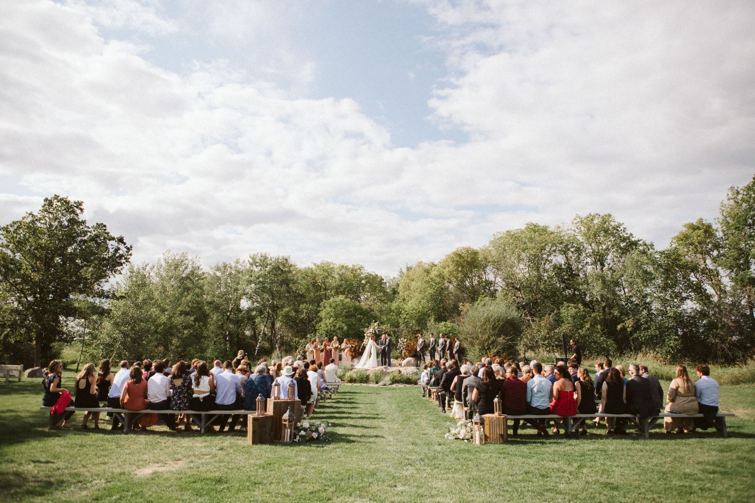 An outdoor wedding ceremony setup with wooden benches and guests on a sunny day surrounded by trees.