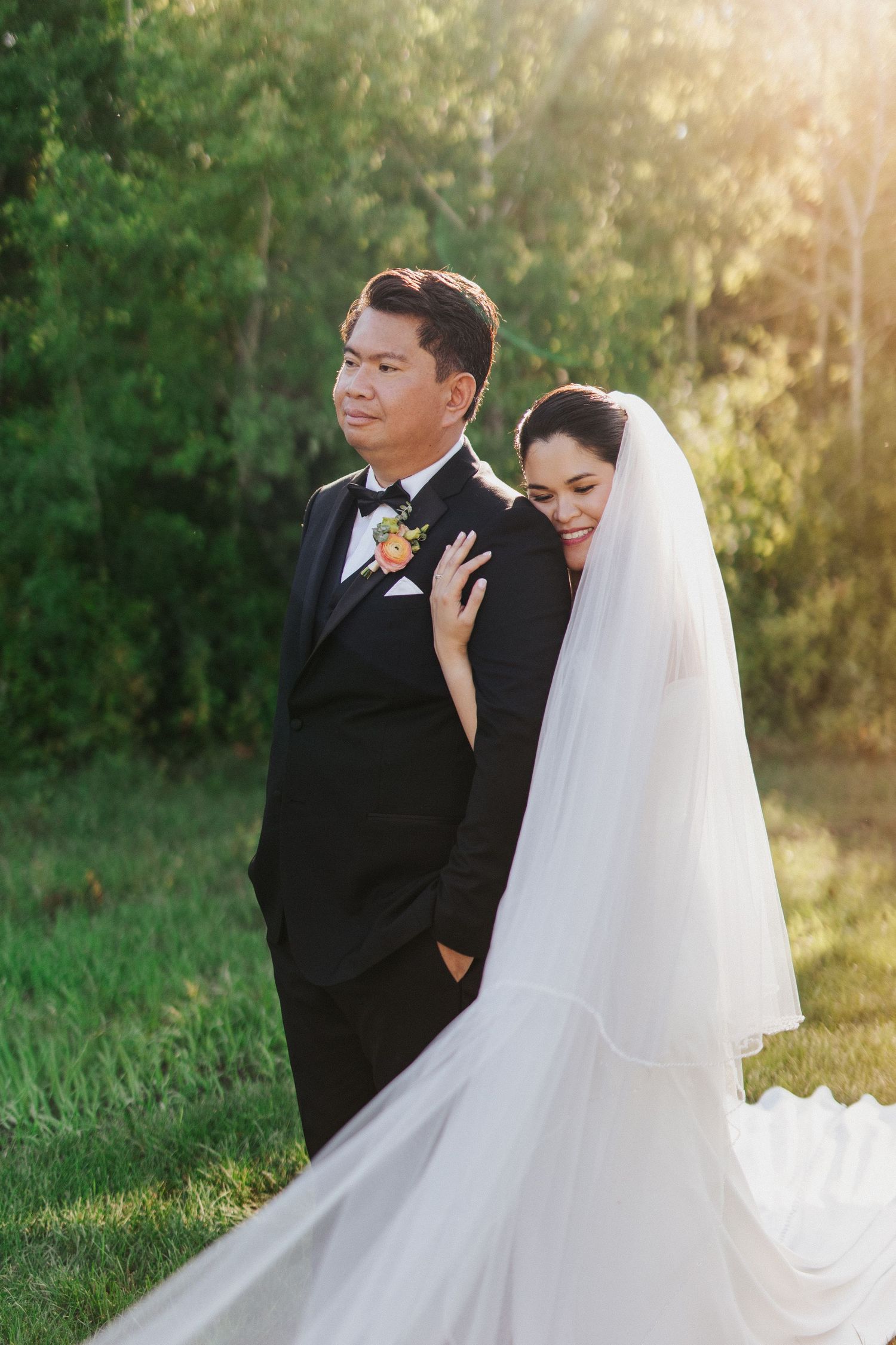Romantic wedding couple poses in golden sunlit field with bride wearing flowing white veil and groom in black suit.