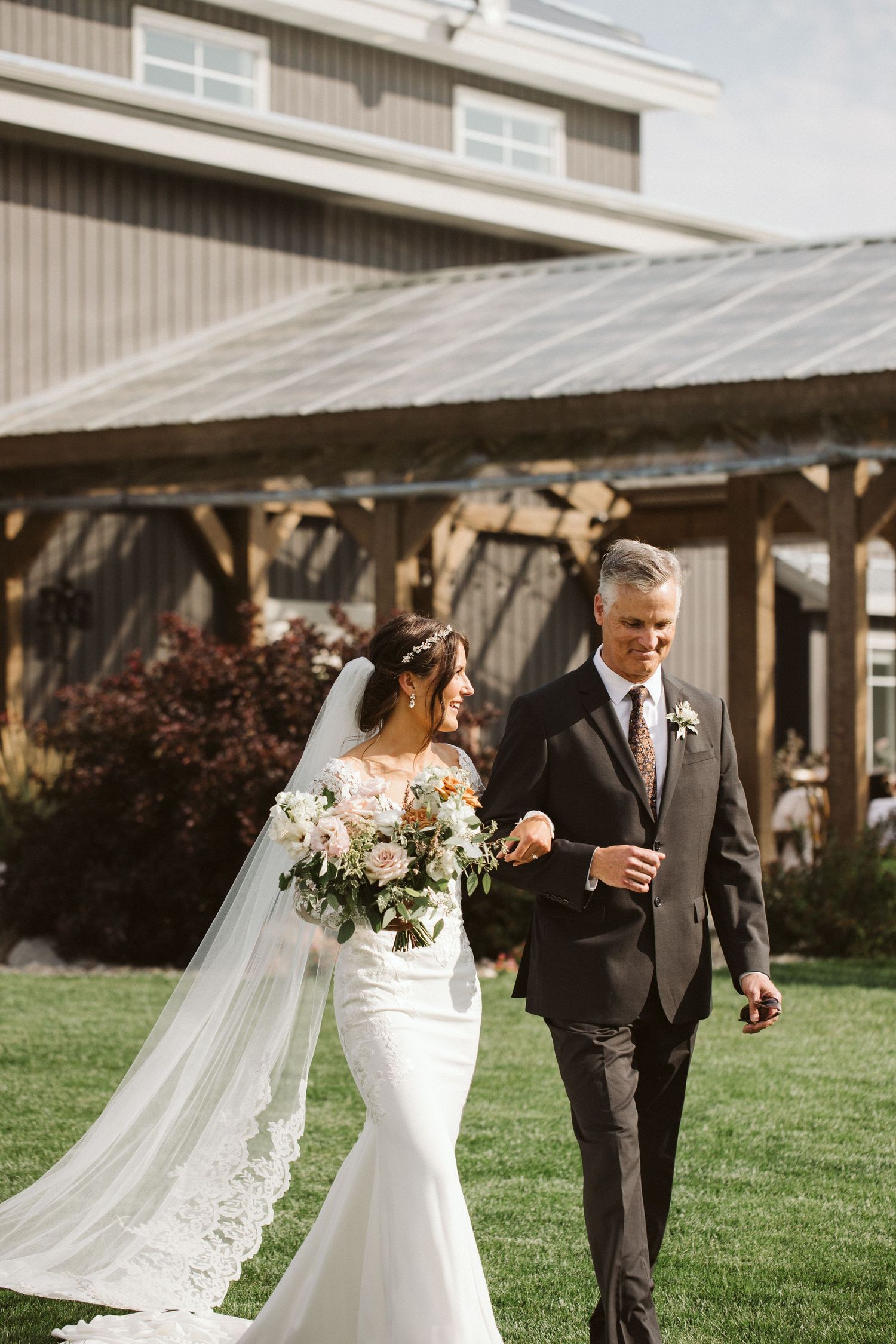Elegant couple walking on green lawn with flowing bridal veil and white bouquet.