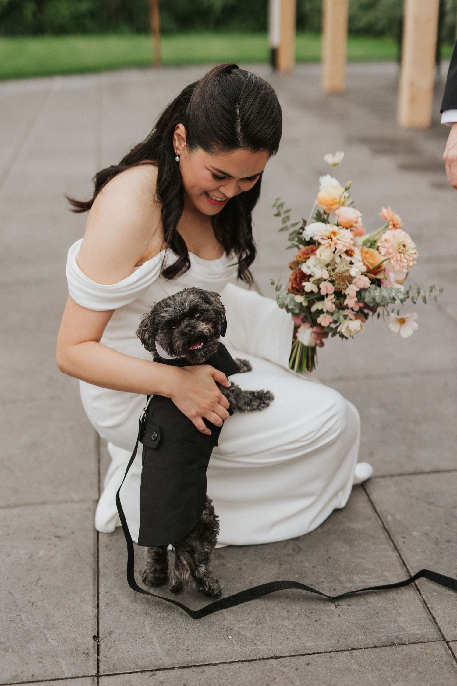 A bride in a white dress kneels down to pet a small black dog on a stone patio with flowers nearby.
