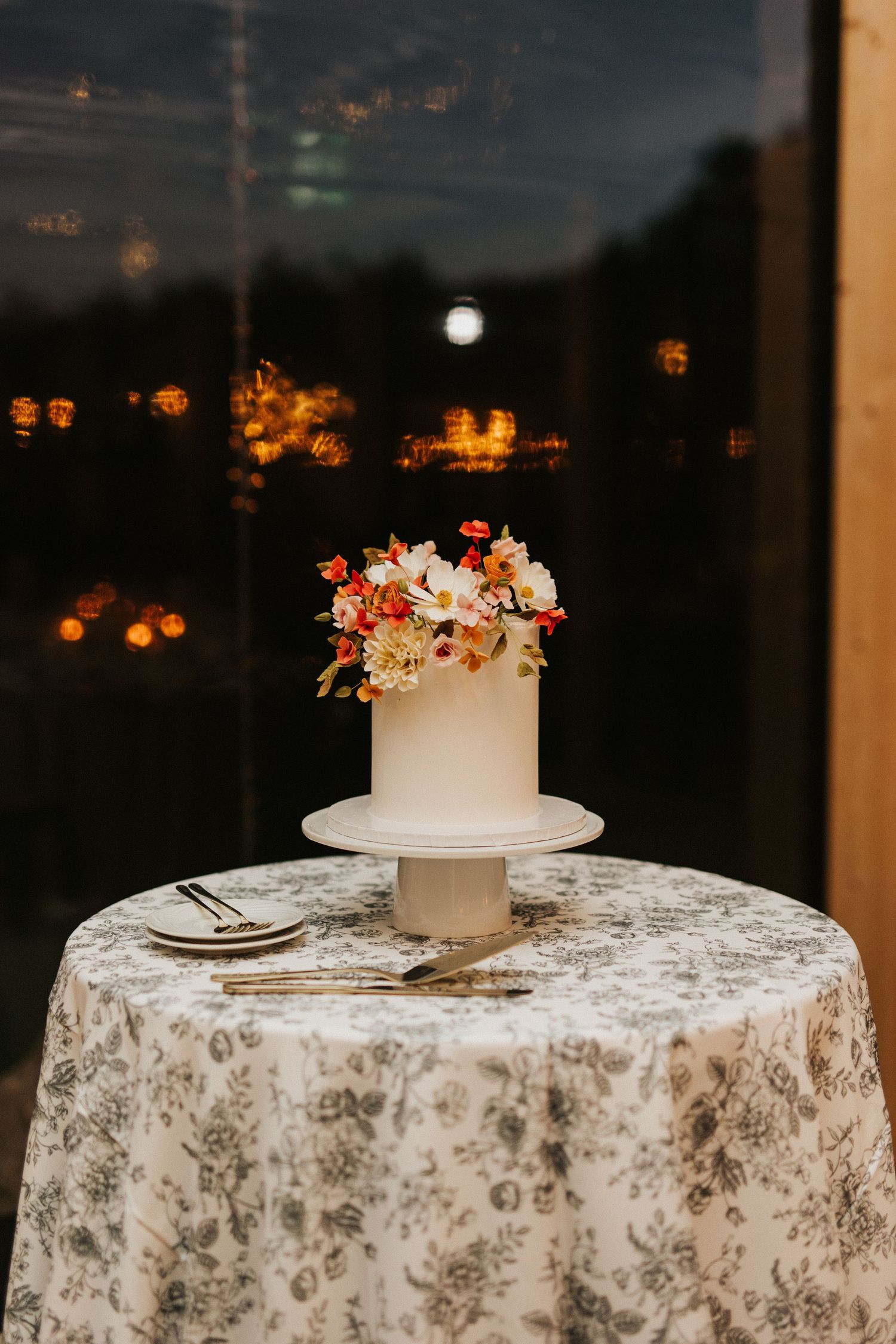 White wedding cakes with floral decorations displayed on patterned tablecloths against a dark window backdrop.