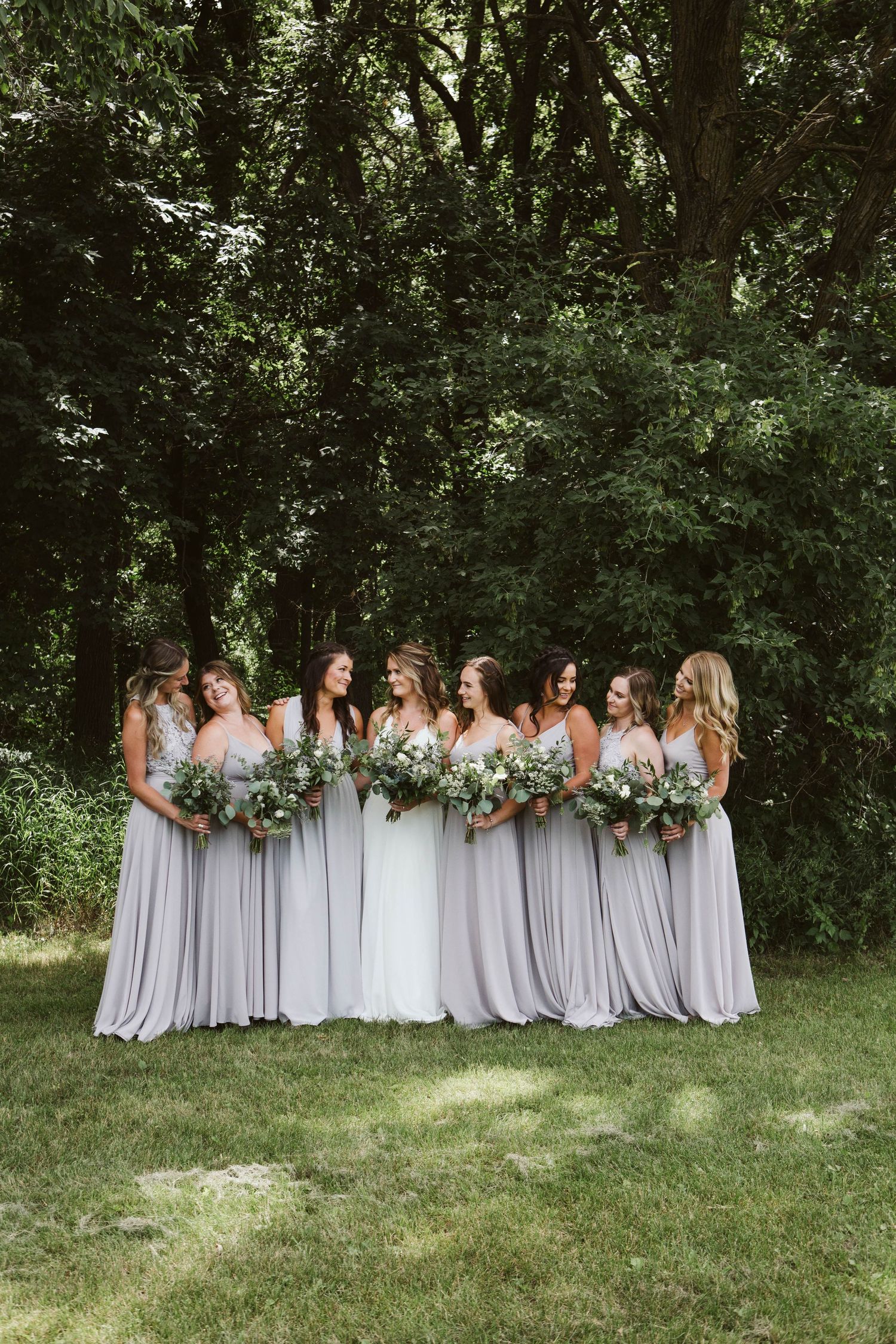 Group of bridesmaids in flowing light grey dresses pose together on grass with floral bouquets.