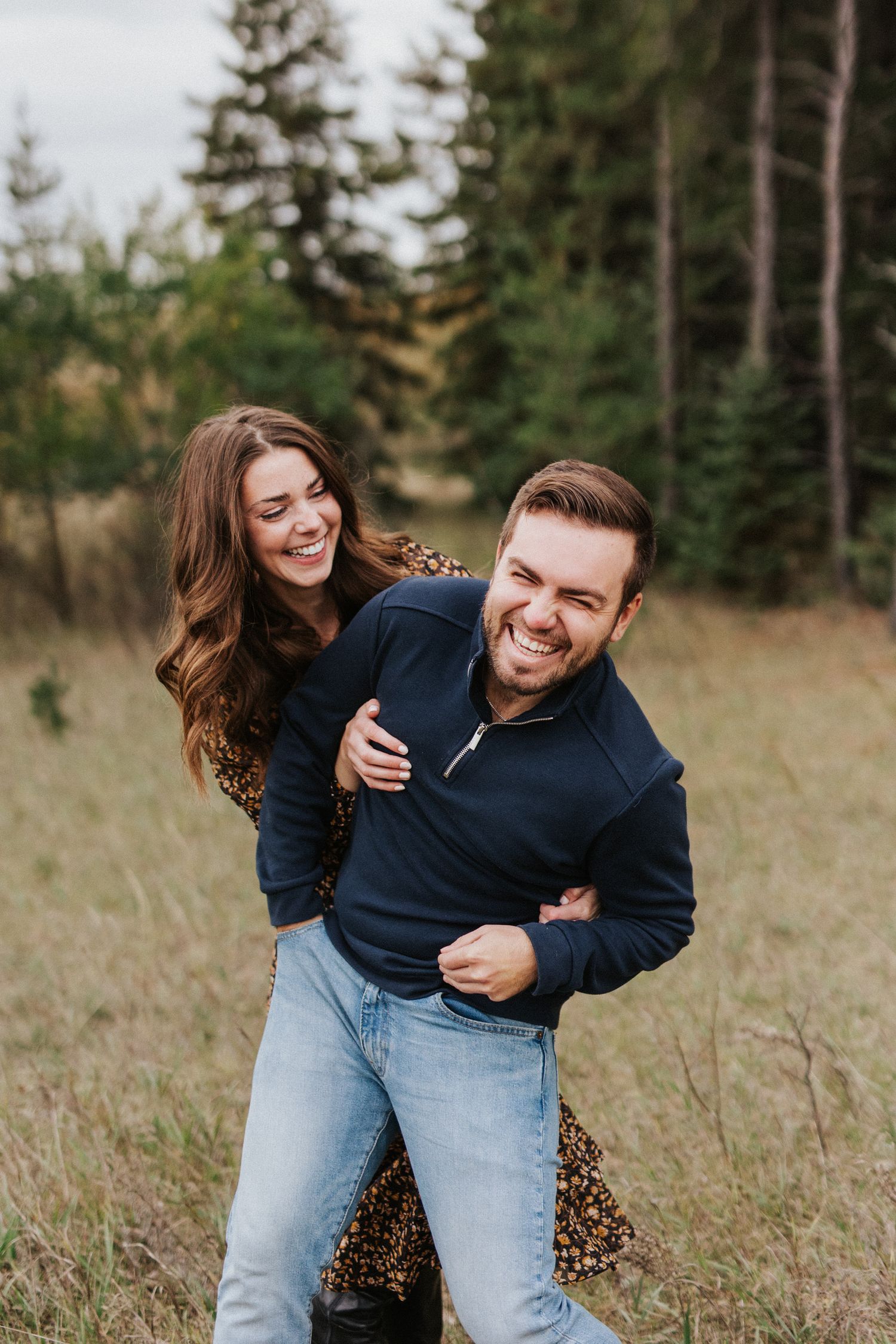 A playful couple laughs together in a grassy field with evergreen trees in the background.