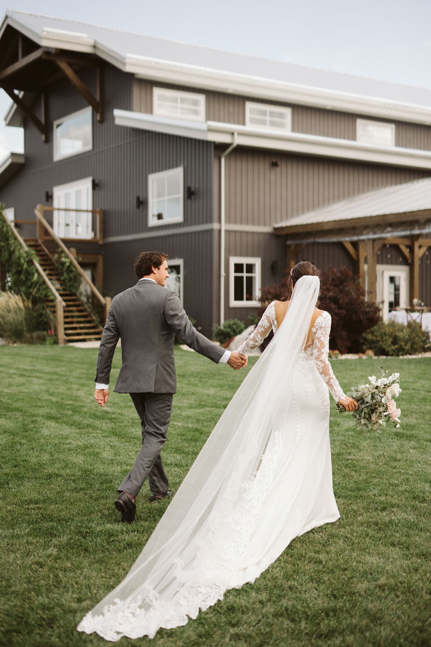 Elegant wedding couple walking on grass in front of a modern barn-style building.