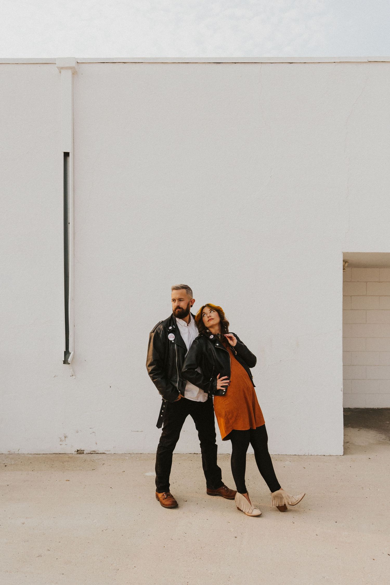 Stylish couple poses against white wall wearing leather jacket and rust-colored dress.