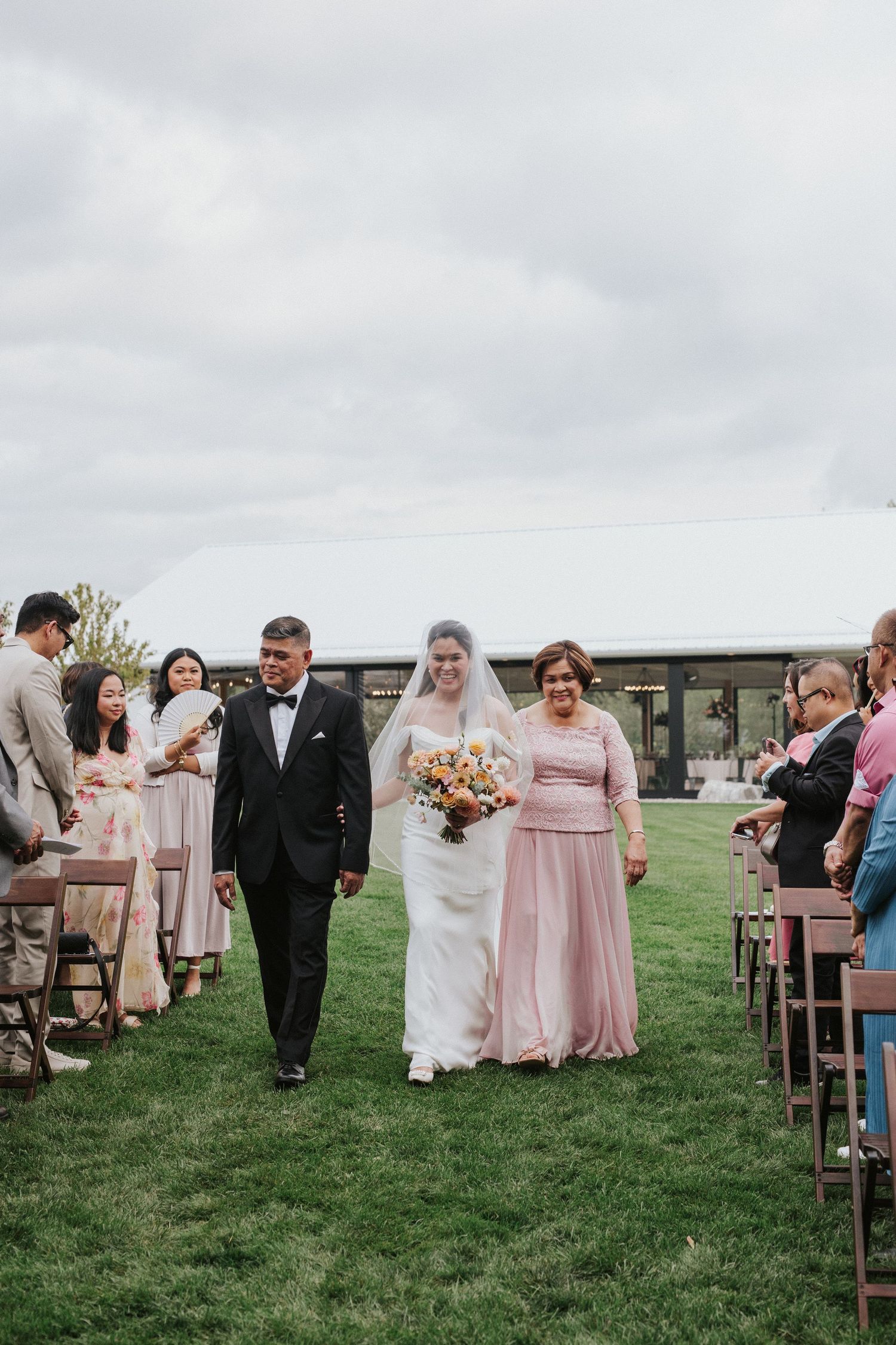 Wedding ceremony guests seated in wooden chairs on a grassy lawn under cloudy skies.