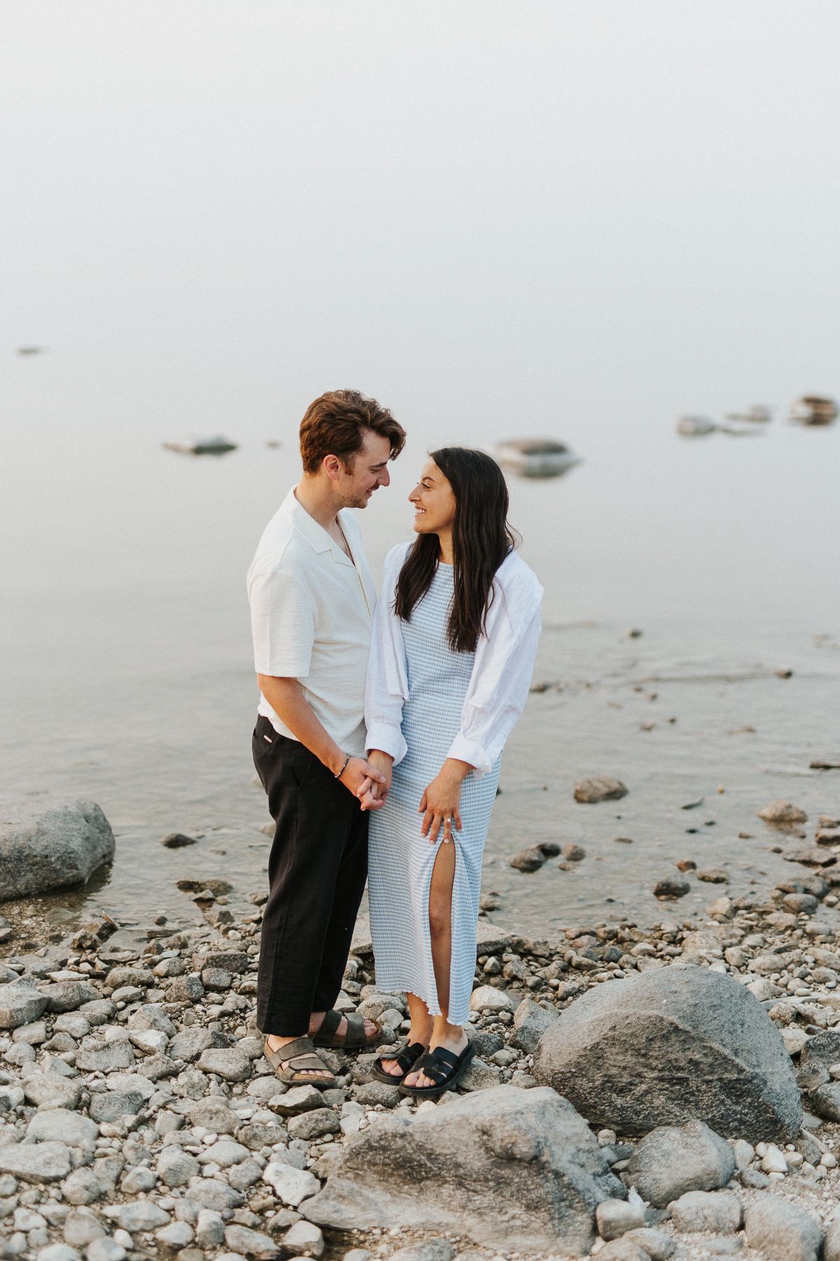 Couple embracing on a rocky shoreline with misty water in background. Gimli, Manitoba, Canada