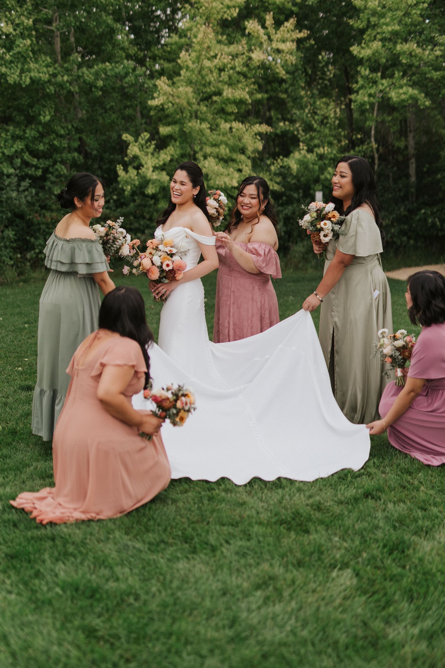 Bridesmaids in sage green and dusty rose dresses arrange the bride's gown on grass during an outdoor photoshoot.