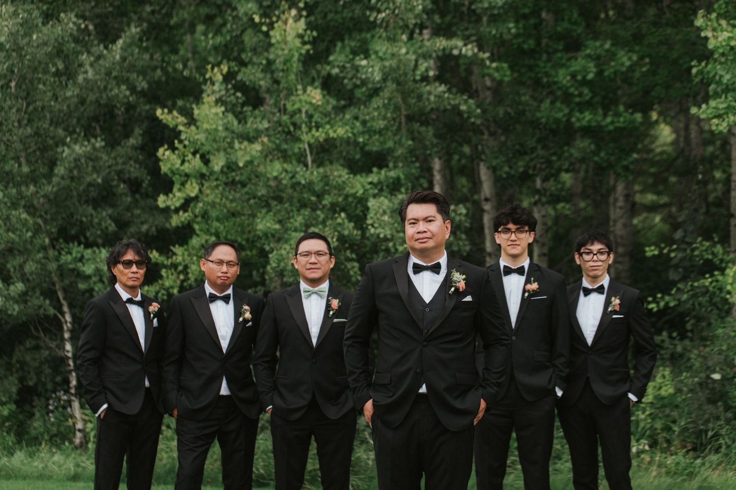 Groomsmen in black suits pose together outdoors in a garden setting for formal wedding photos.