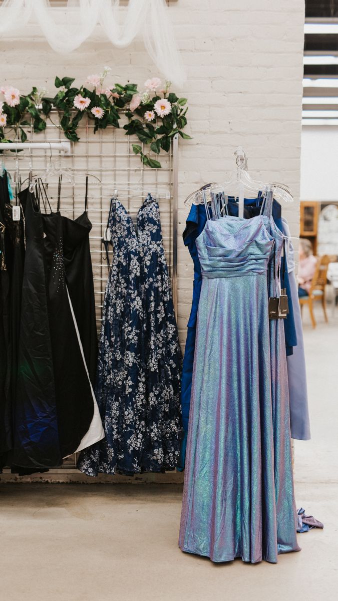 Blue and floral formal dresses displayed on hangers against a white wall.