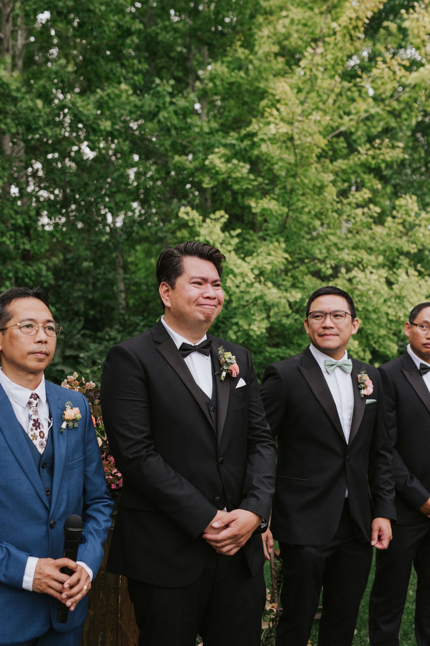 Groomsmen in dark suits and ties stand in line against a leafy green backdrop.