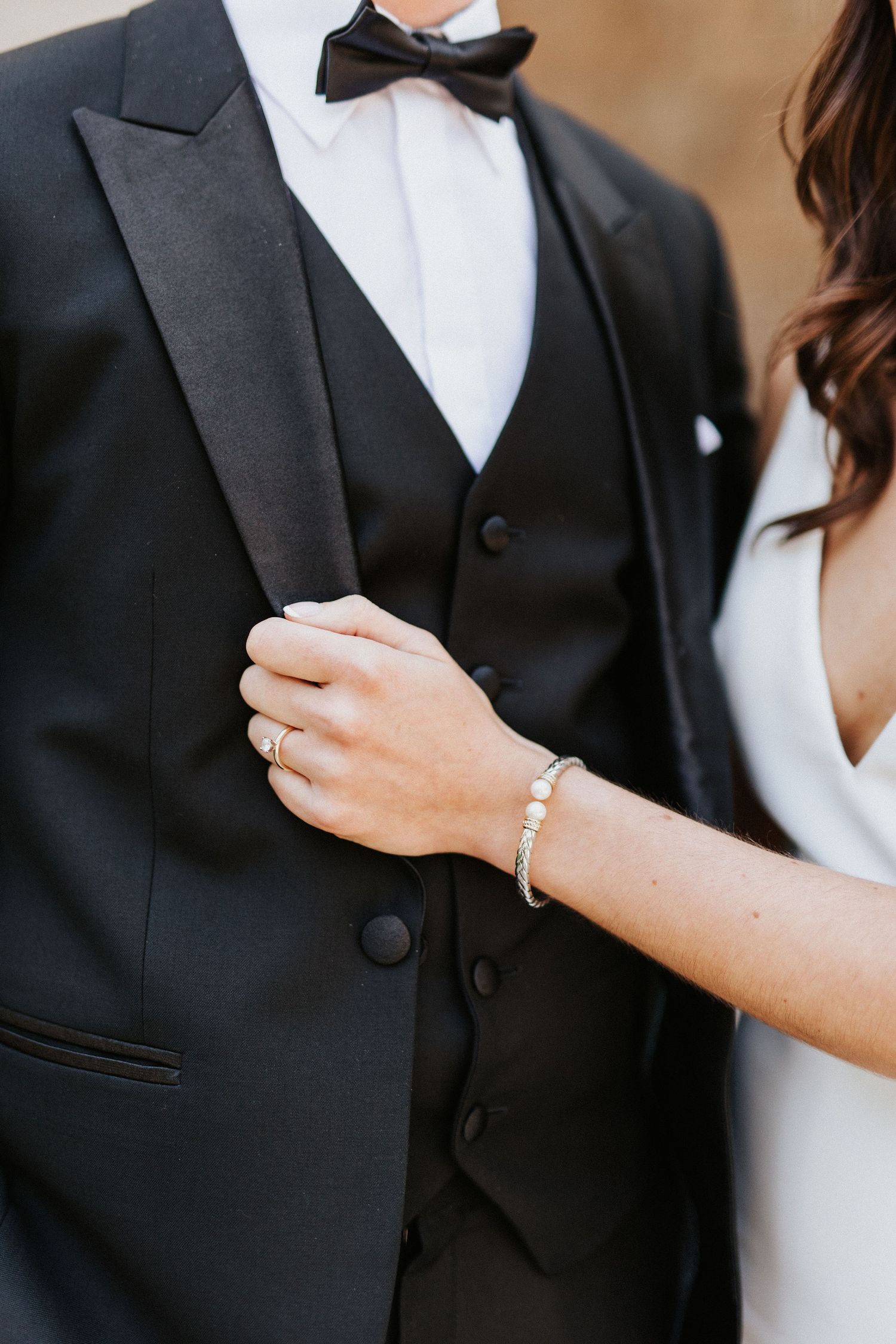 Close-up details of formal wedding attire showing rings, jewelry, and black tuxedo.