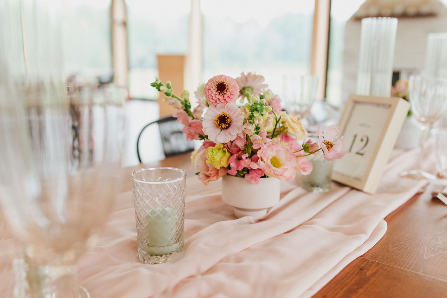 Elegant wedding table setting with pink floral centerpiece, glass, and table number in soft natural light.