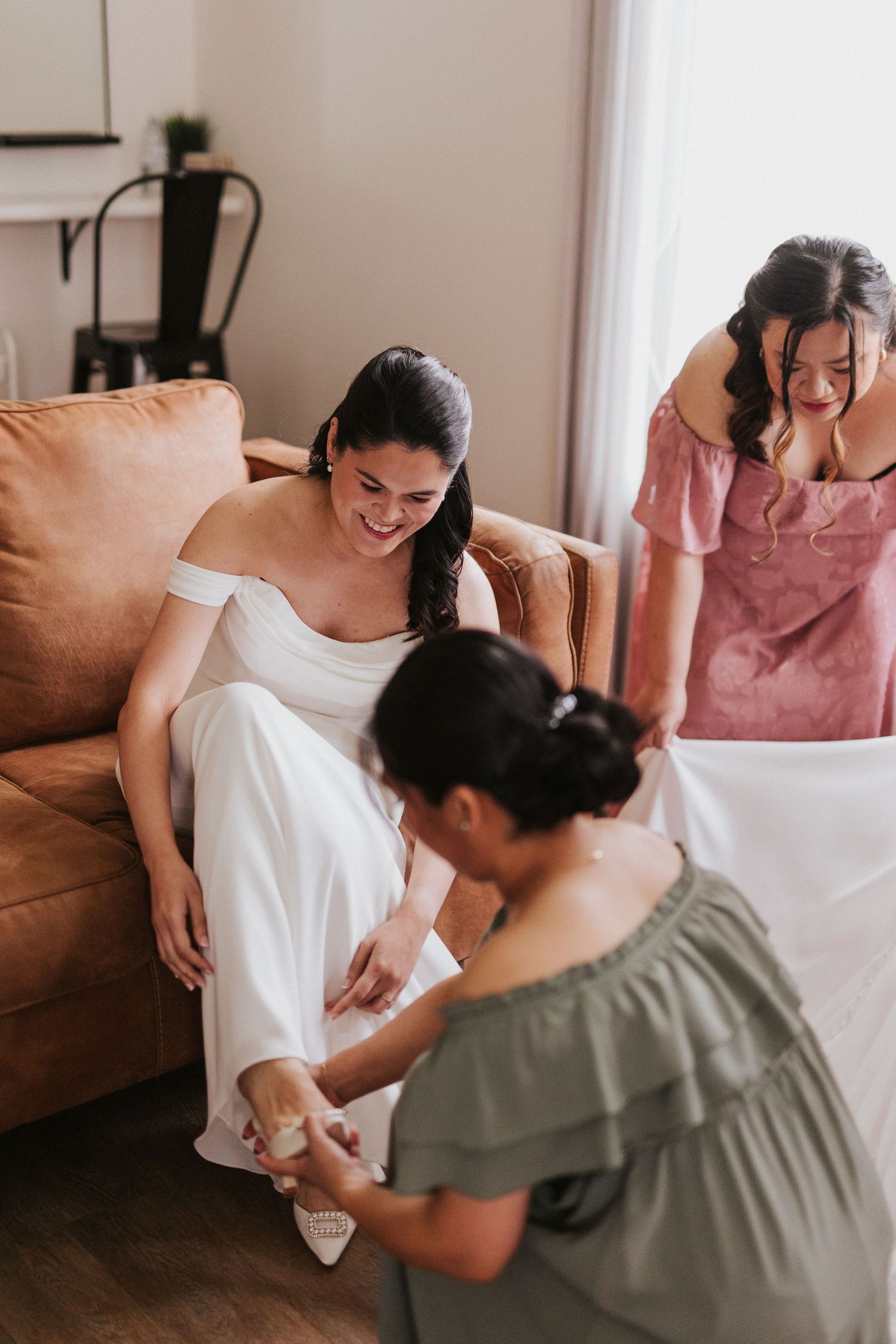 Wedding party members help the bride put on her shoes while sitting on a tan couch in a sunlit room.