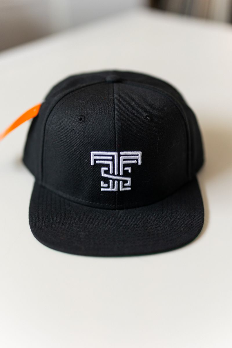 Close-up of black cap with white geometric embroidered logo on flat surface.