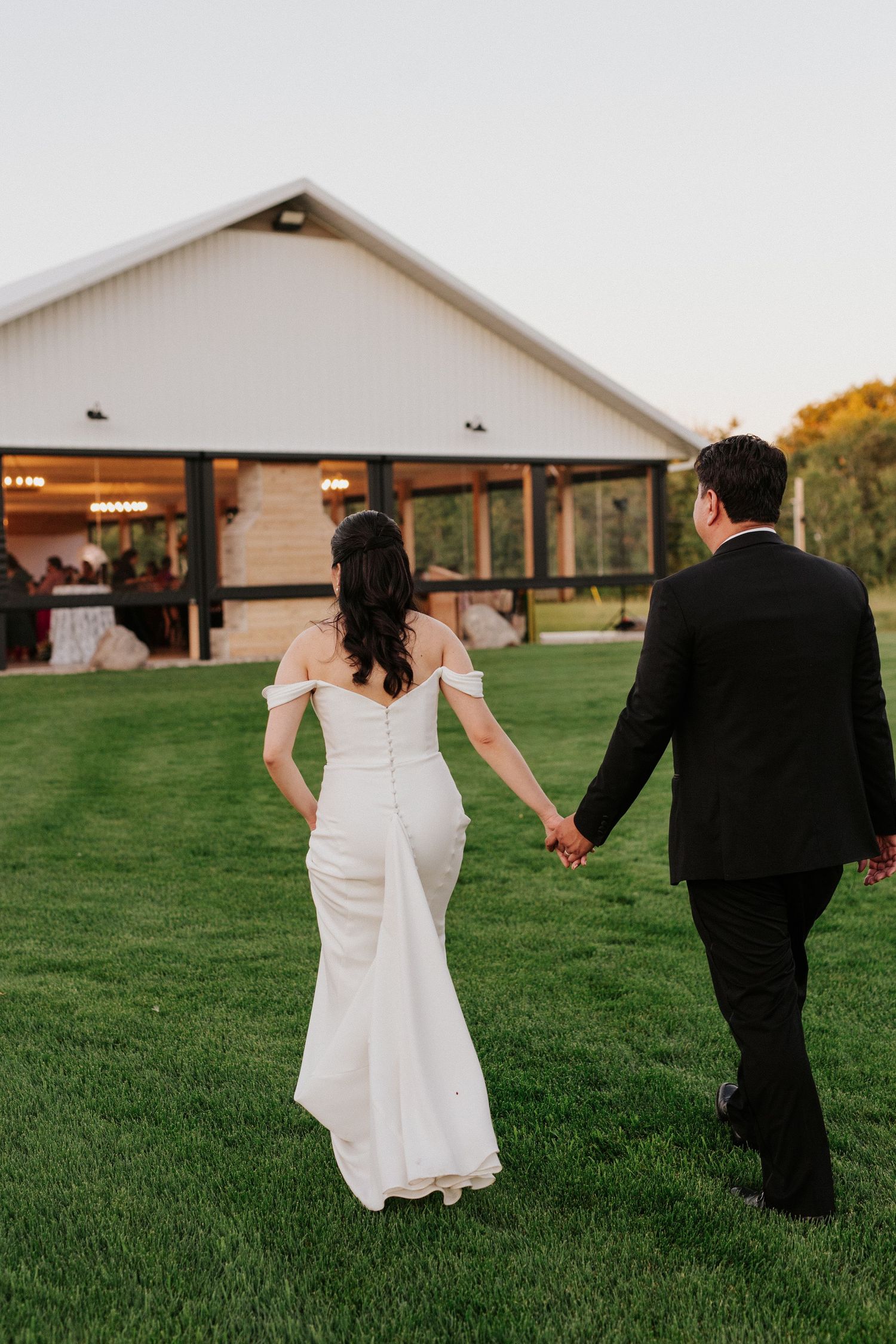 A couple in wedding attire walks away together across a lush green lawn at sunset.